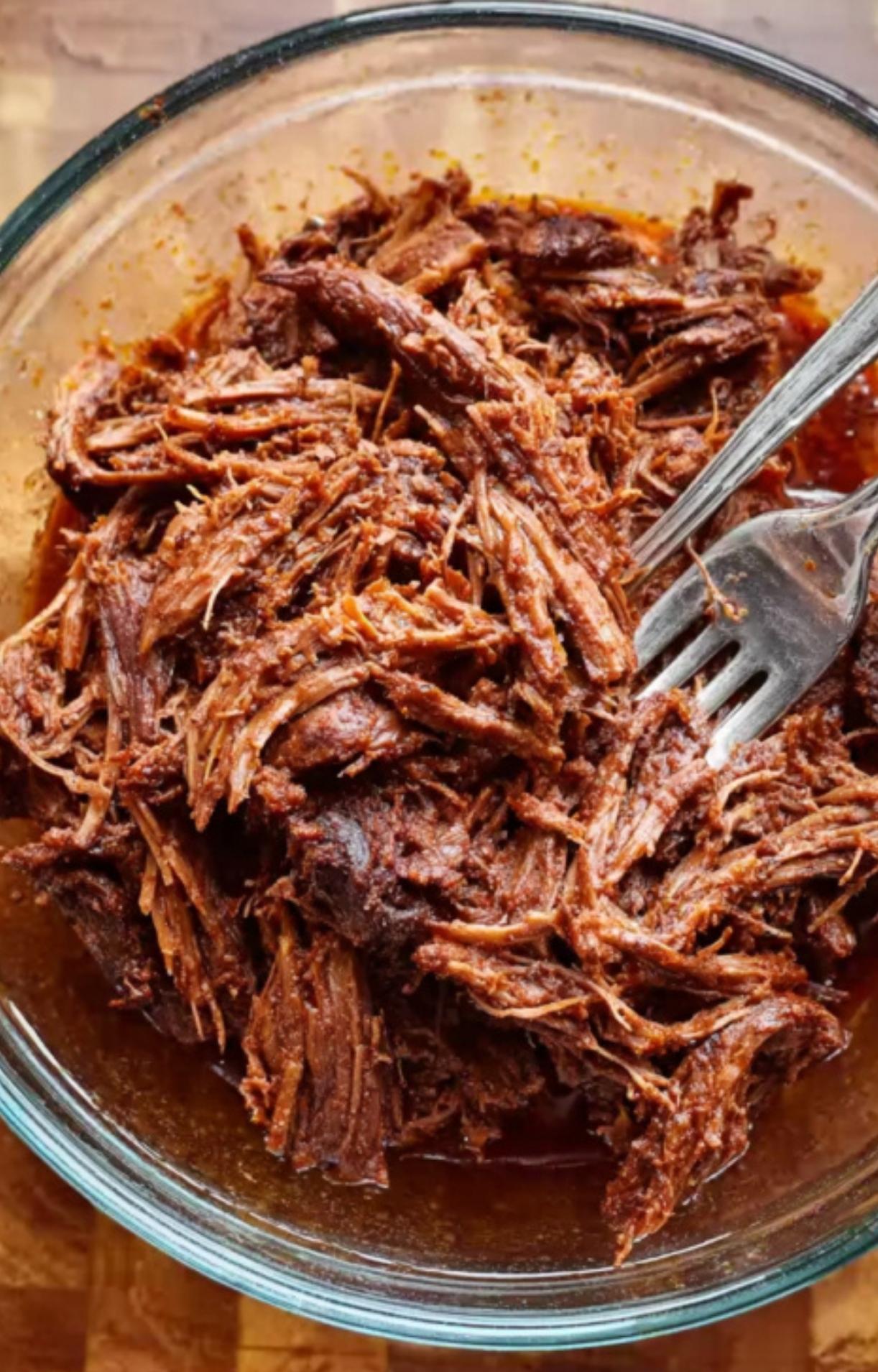 Tender, slow-cooked beef being shredded with two silver forks in a glass bowl filled with a bit of the flavorful cooking liquid.