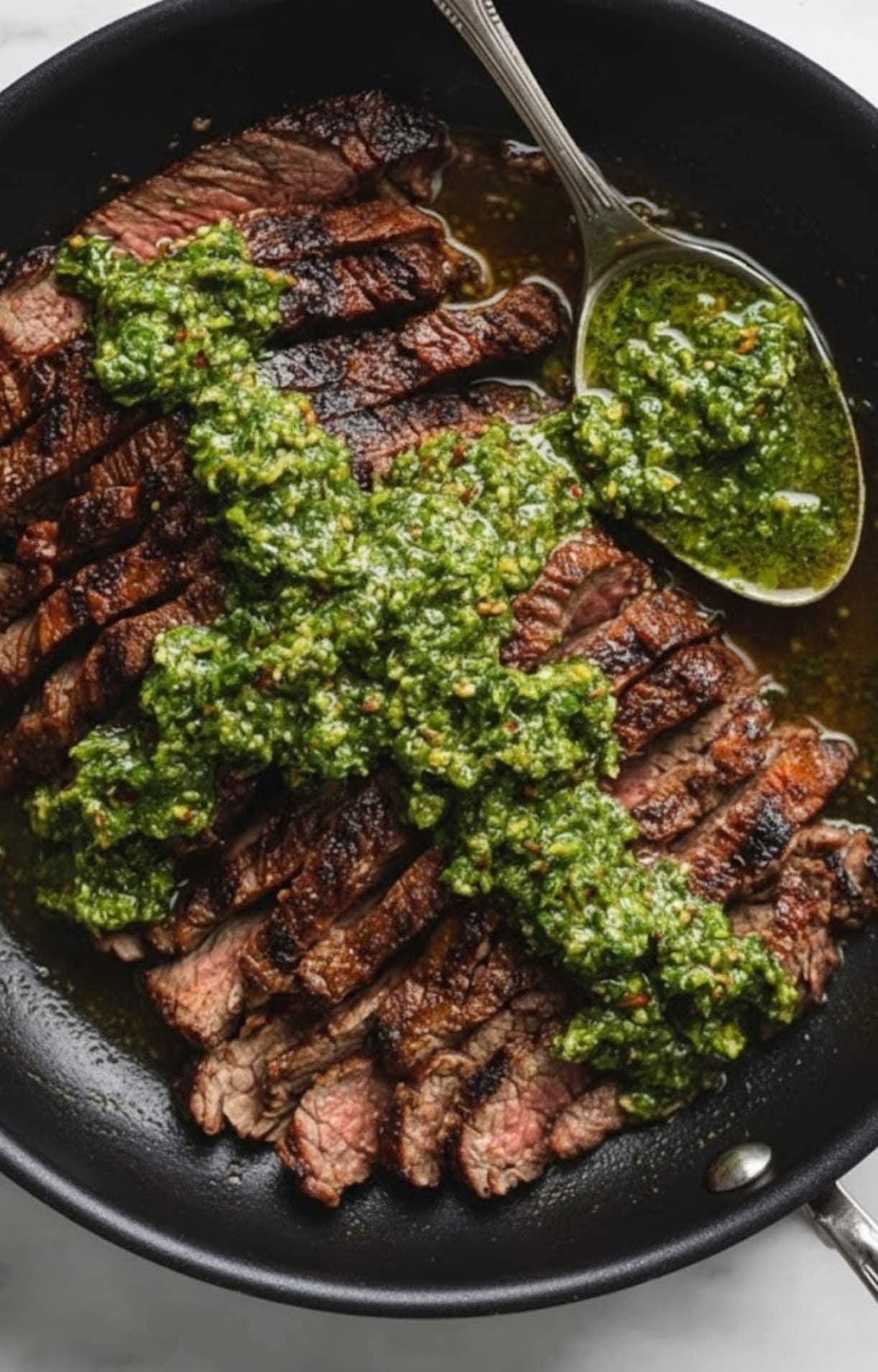 featuring a close-up of a silver spoon resting in a dark skillet alongside expertly charred, medium-rare steak slices that are being layered with a vibrant, herb-heavy green sauce.