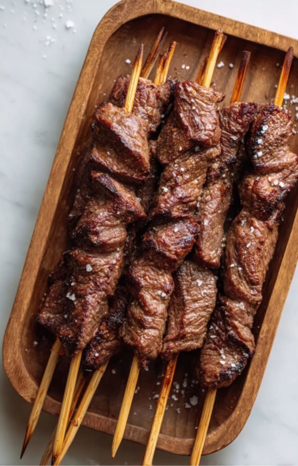 Wooden tray filled with hot, grilled steak skewers sprinkled with flaky sea salt.