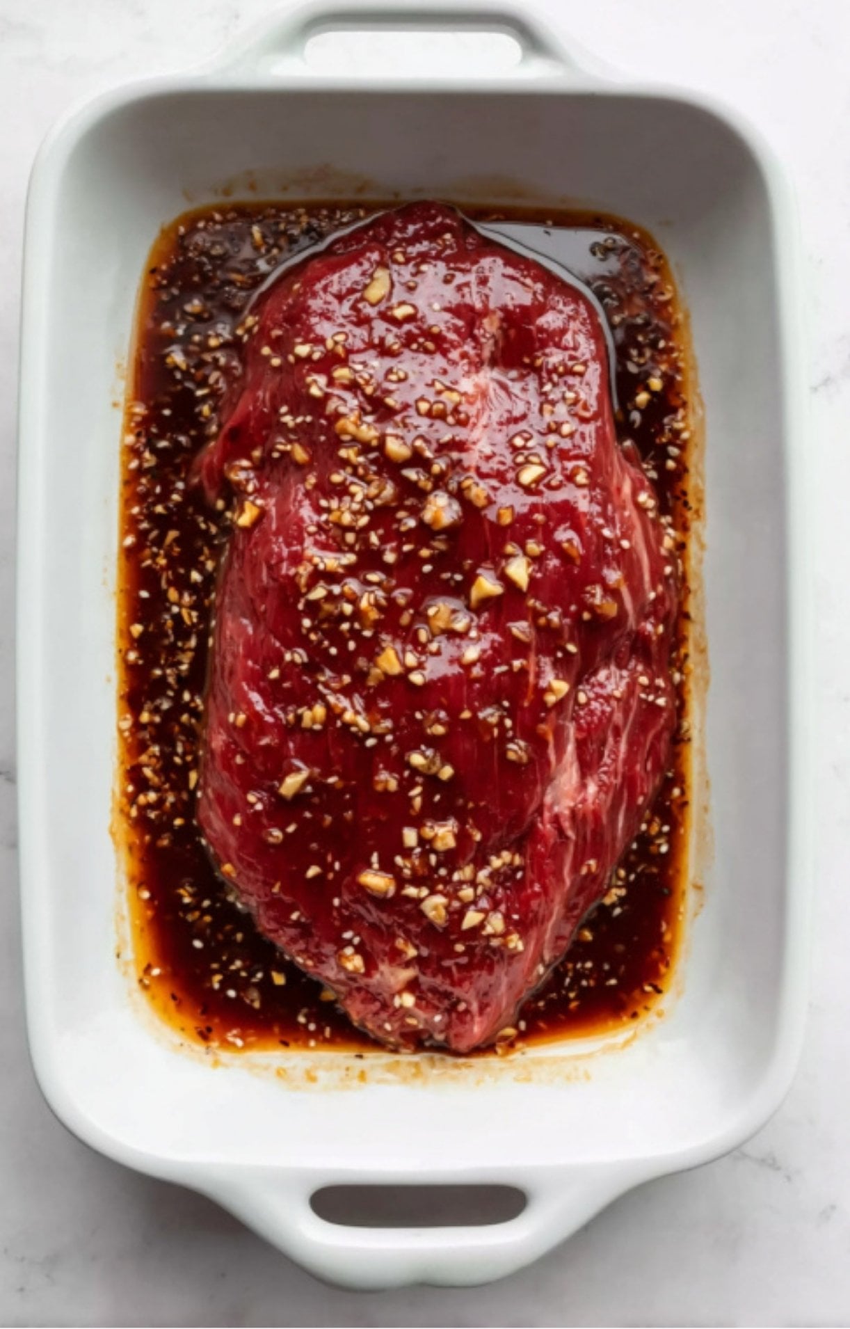 Raw flank steak soaking in a garlic and soy marinade in a white baking dish, ready for grilling.