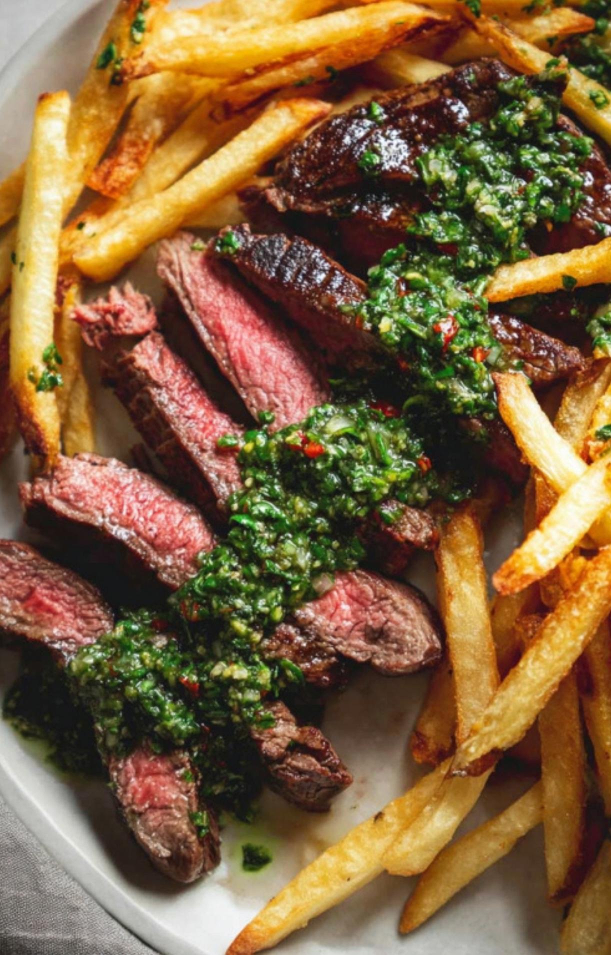 Sliced steak and crispy fries on a plate, drizzled with green chimichurri sauce.