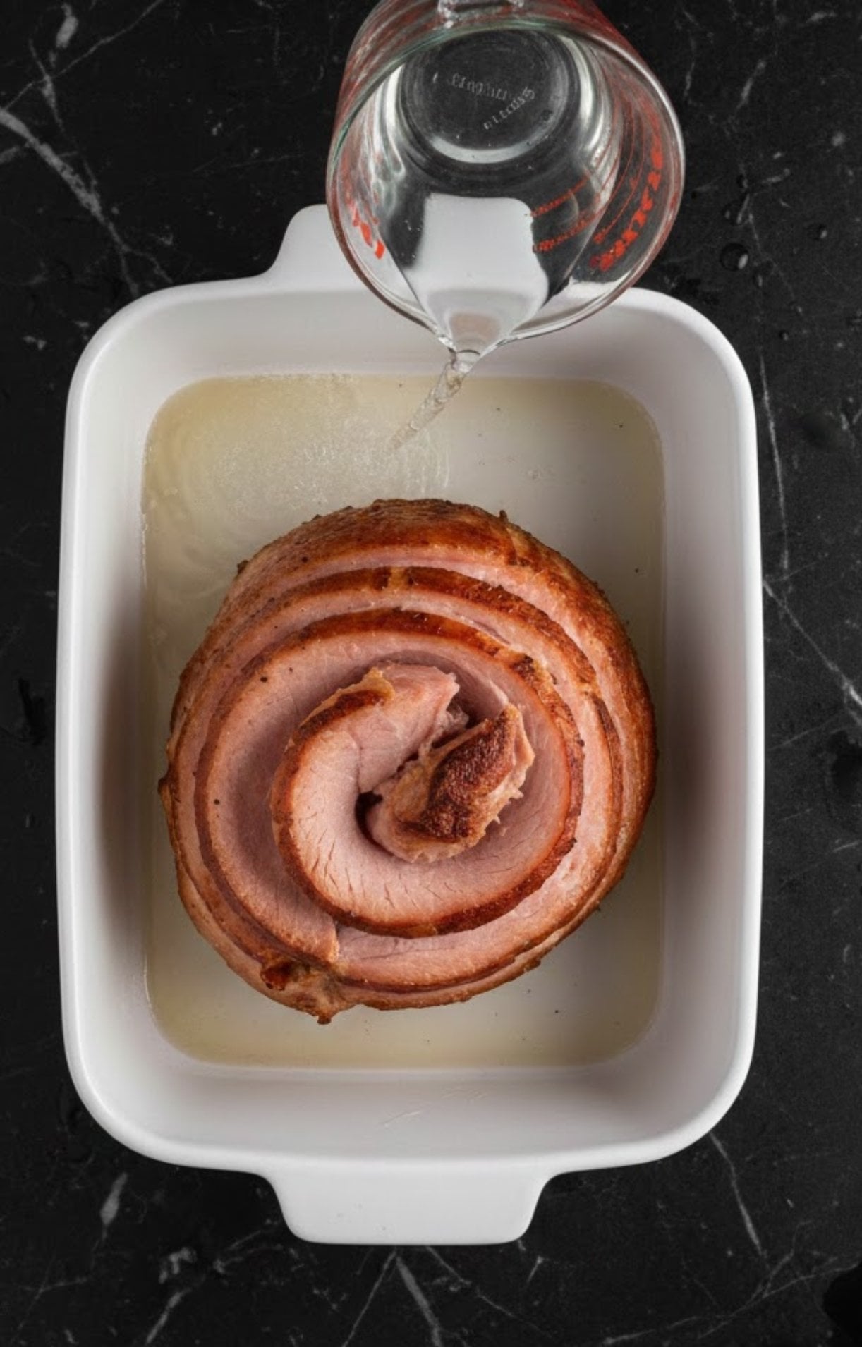 Pouring water into baking dish with smoked ham, steam keeps ham juicy while reheating