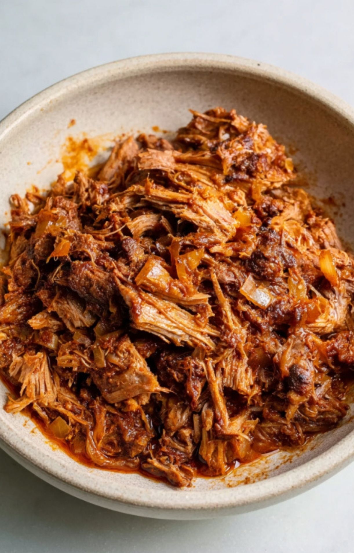A ceramic bowl filled with tender, slow-cooked shredded beef coated in a rich, reddish-brown chili sauce.