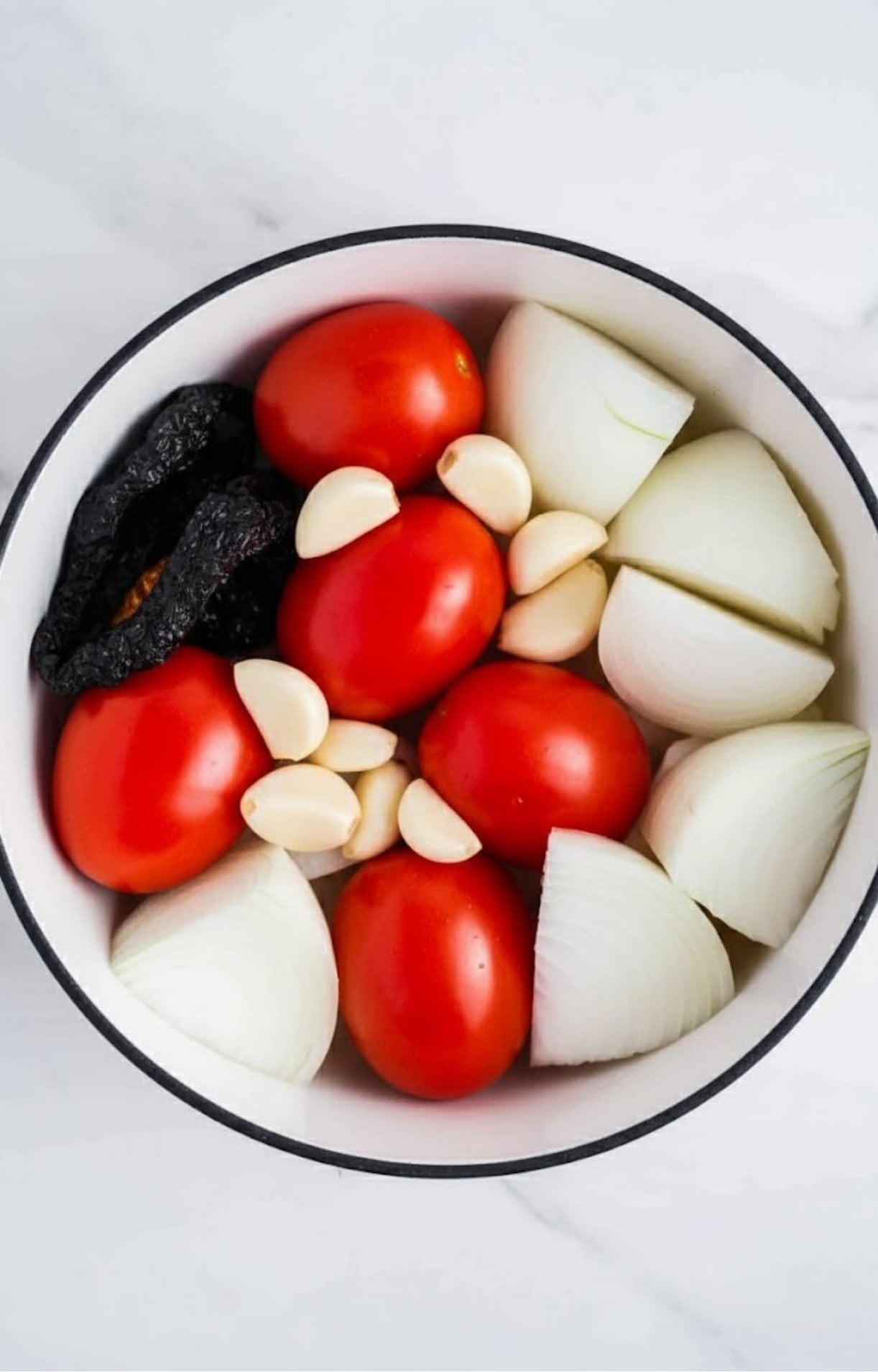 Raw Roma tomatoes, quartered white onions, garlic cloves, and dried guajillo chilies in a white pot.