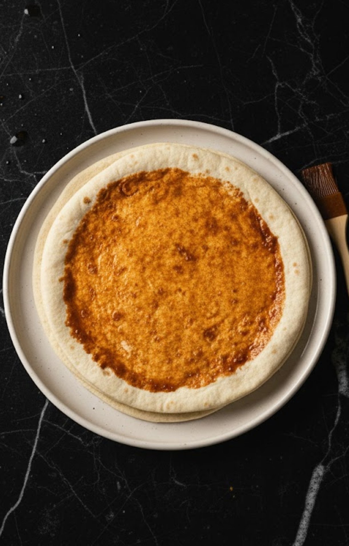 A pastry brush applying a layer of reddish birria oil or sauce to a round pizza dough.
