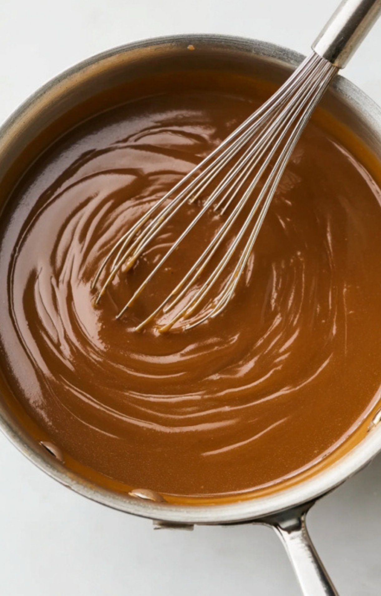 A stainless steel saucepan filled with a smooth, glossy brown gravy being stirred with a wire whisk.