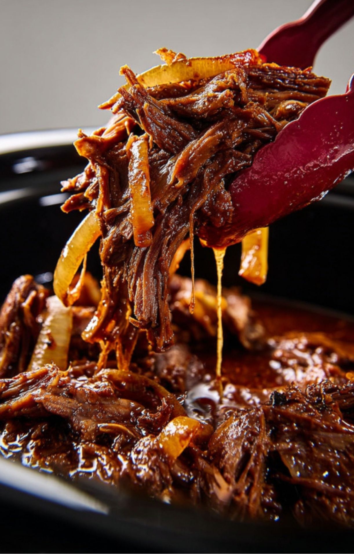A pair of red kitchen tongs lifting a heap of tender, succulent shredded beef and cooked onions from a slow cooker.