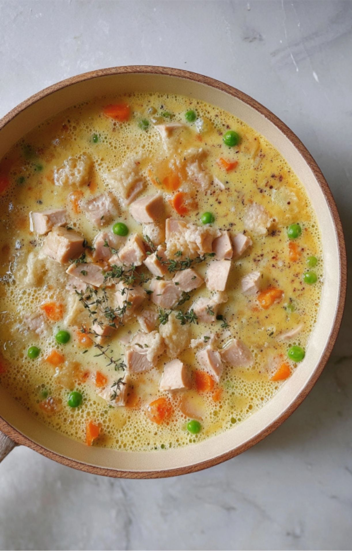 Overhead view of turkey pot pie filling simmering in a skillet with diced turkey, peas, carrots, and fresh thyme in a creamy yellow broth.