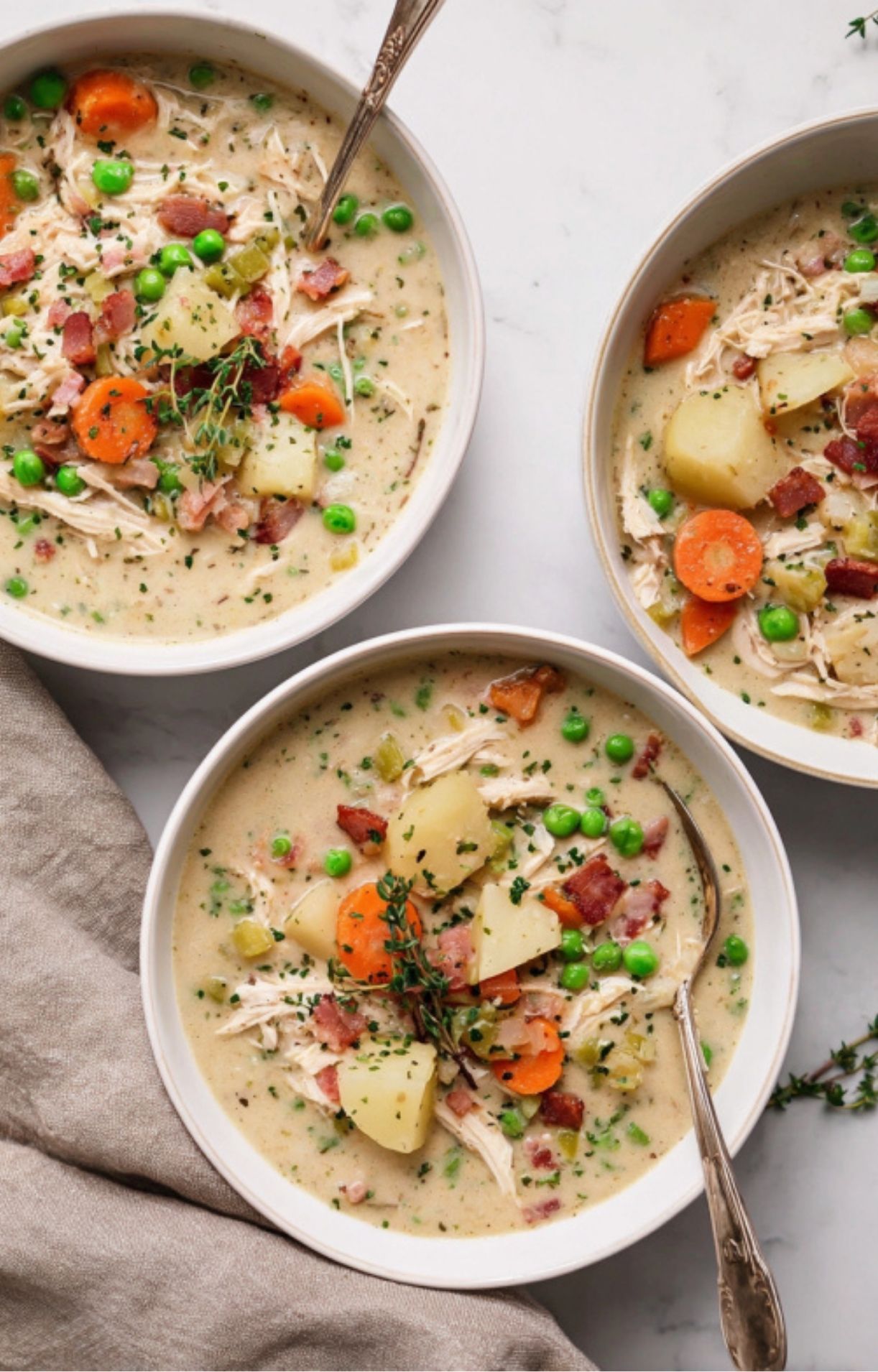 Three white bowls of Turkey Pot Pie Soup arranged on a marble surface, each garnished with a sprig of fresh thyme and bacon.