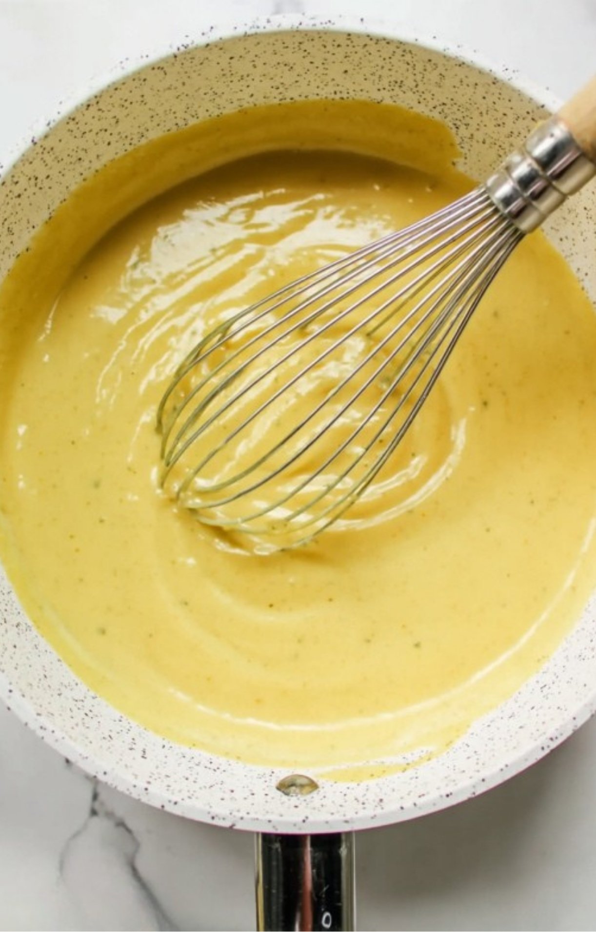 A metal whisk stirring a thick, smooth yellow gravy in a white speckled saucepan.