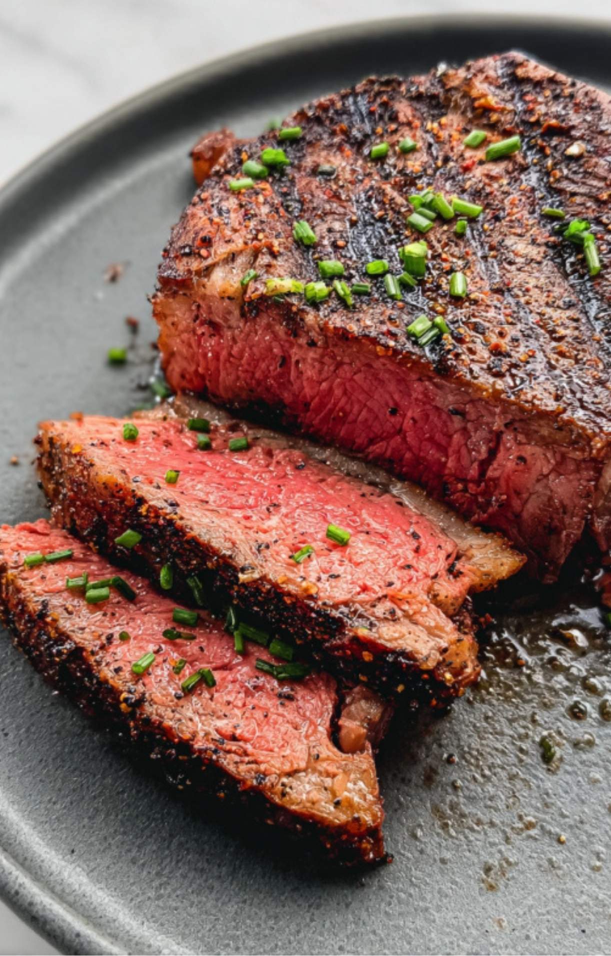 A close-up of a grilled ribeye steak sliced to reveal a tender, pink medium-rare center, seasoned with a herb-flecked dry rub and garnished with chives.