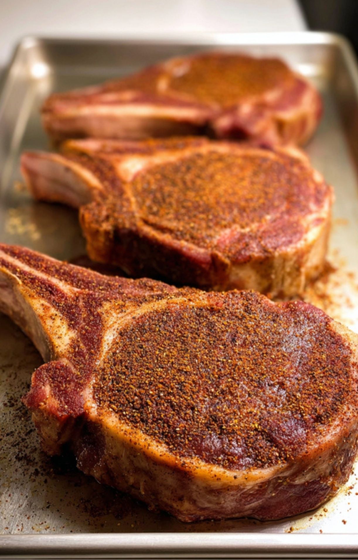 Three bone-in rib eye steaks fully covered in a dark, savory spice rub, resting on a sheet pan before heading to the grill or pan.