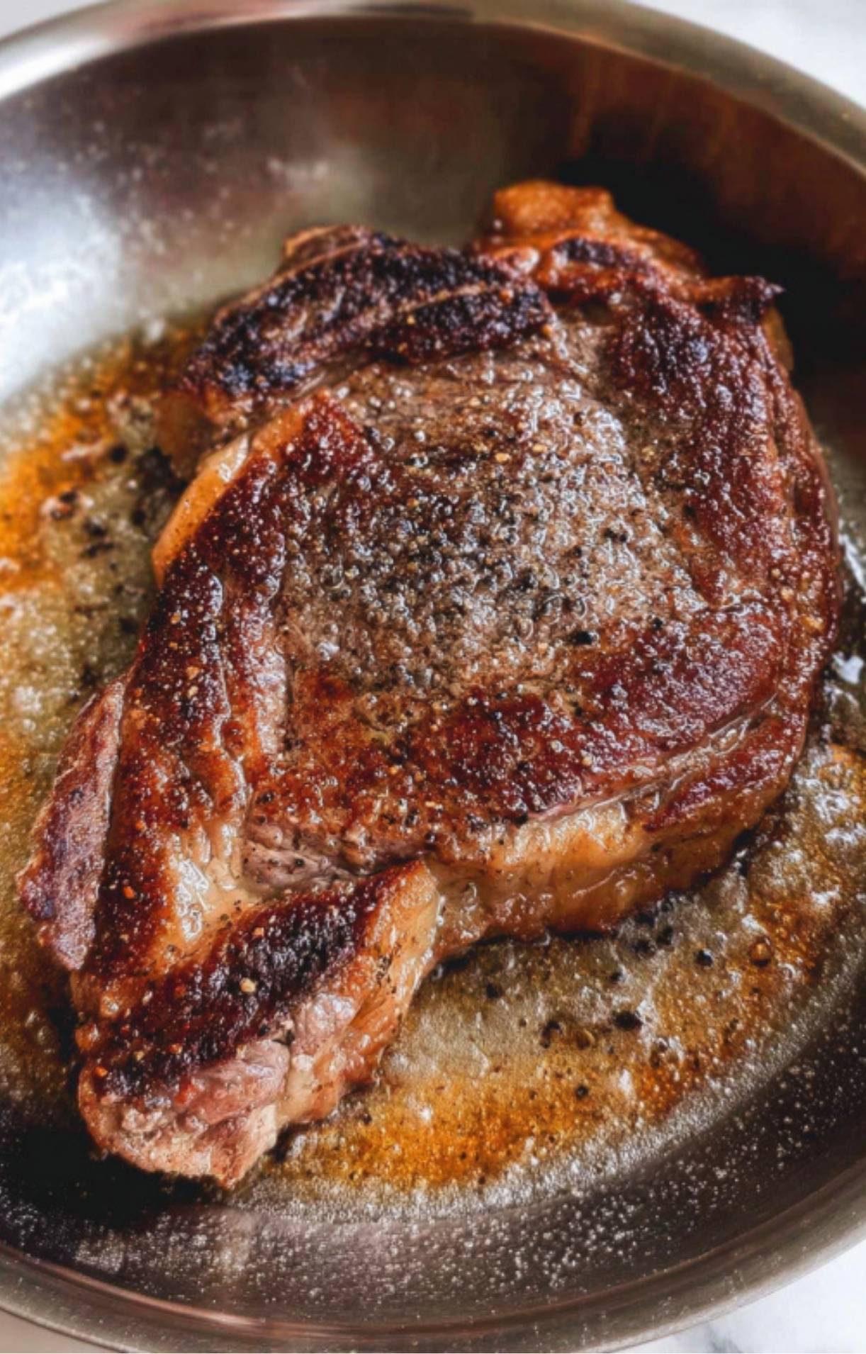 A thick ribeye steak searing in a stainless steel pan with bubbling butter and black pepper.