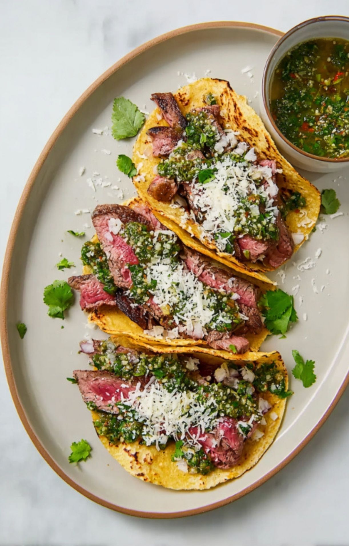 Three grilled steak tacos topped with bright green cilantro chimichurri, shredded cheese, and fresh cilantro, served on a plate with a bowl of chimichurri sauce.