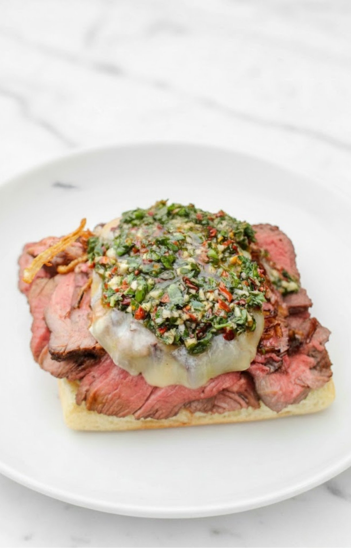 Open-faced sandwich with sliced grilled steak, melted provolone cheese, and a generous spoonful of chimichurri sauce on toasted bread.