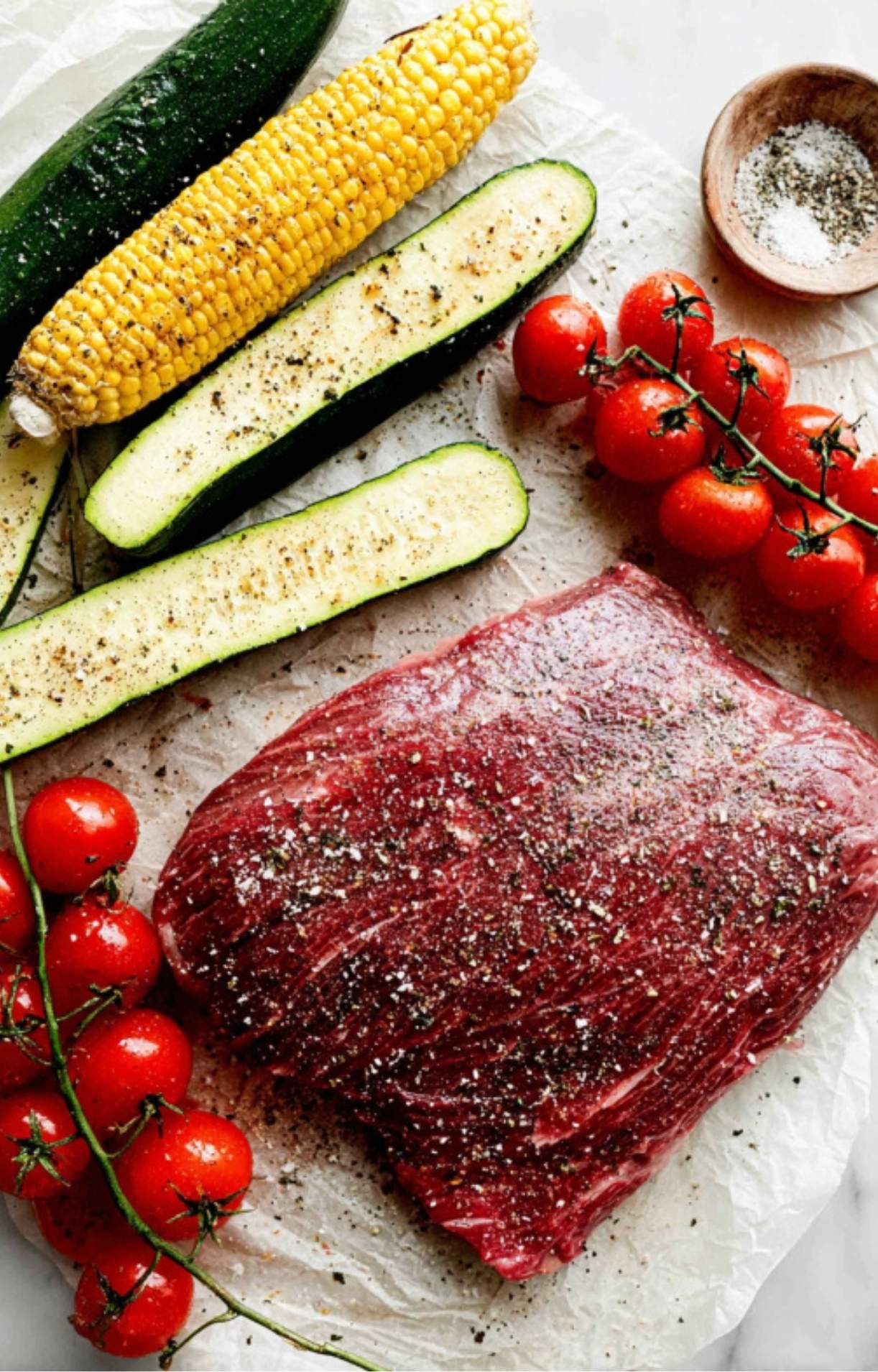 Raw flank steak, zucchini, corn on the cob, and cherry tomatoes sprinkled with salt and pepper, ready to be grilled