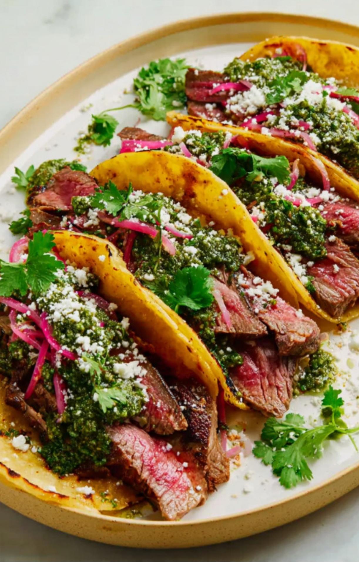 A serving plate with several grilled steak tacos, loaded with green chimichurri, pickled onions, cotija cheese, and sprigs of cilantro.