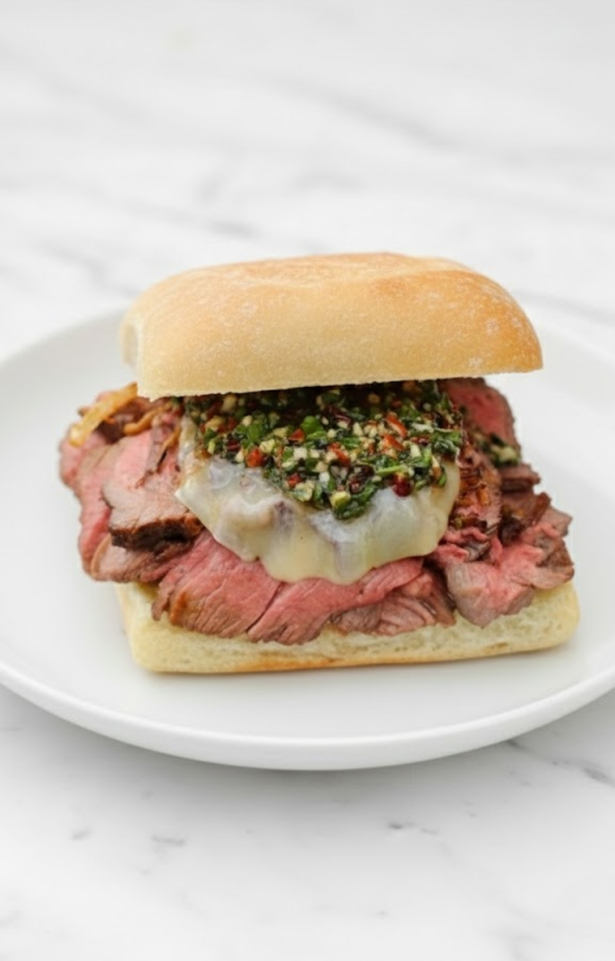 A grilled steak sandwich on a ciabatta roll, layered with juicy sliced steak, melted provolone cheese, crispy fried onions, and a generous spoonful of fresh chimichurri sauce.