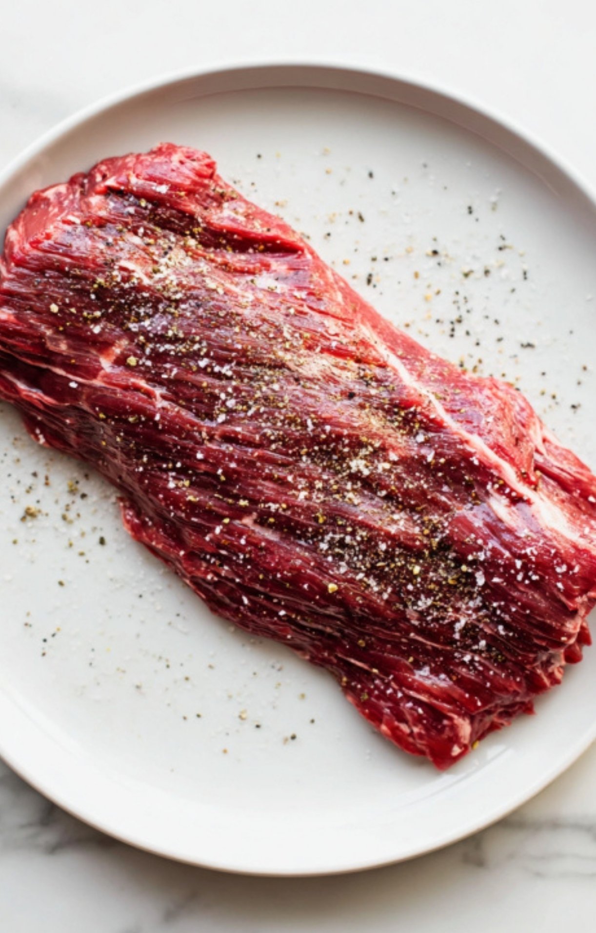 Uncooked skirt steak seasoned with salt and black pepper on a round white plate, prepared for the grill.