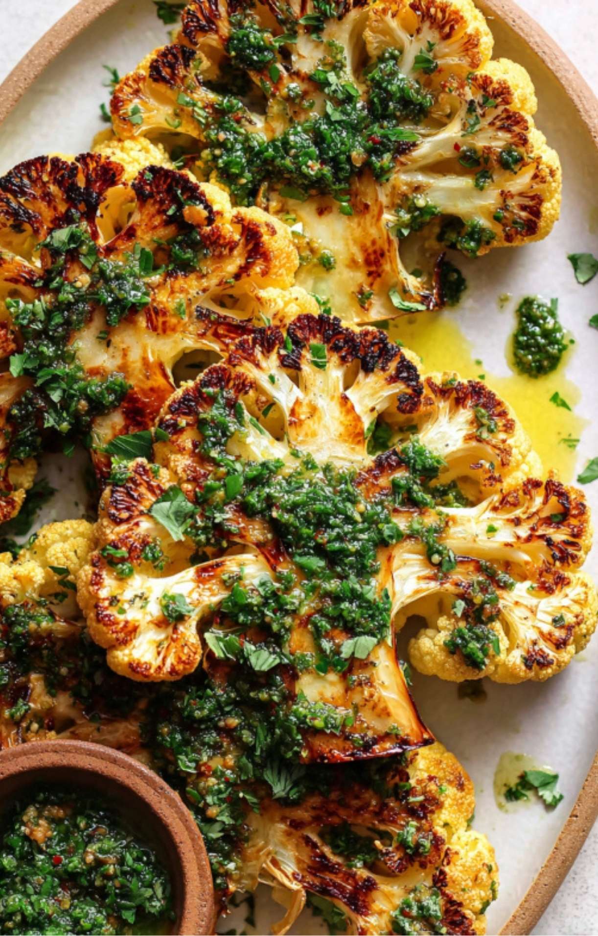 Crispy, golden-brown grilled cauliflower steaks on a platter, topped with vibrant green chimichurri sauce and fresh parsley.