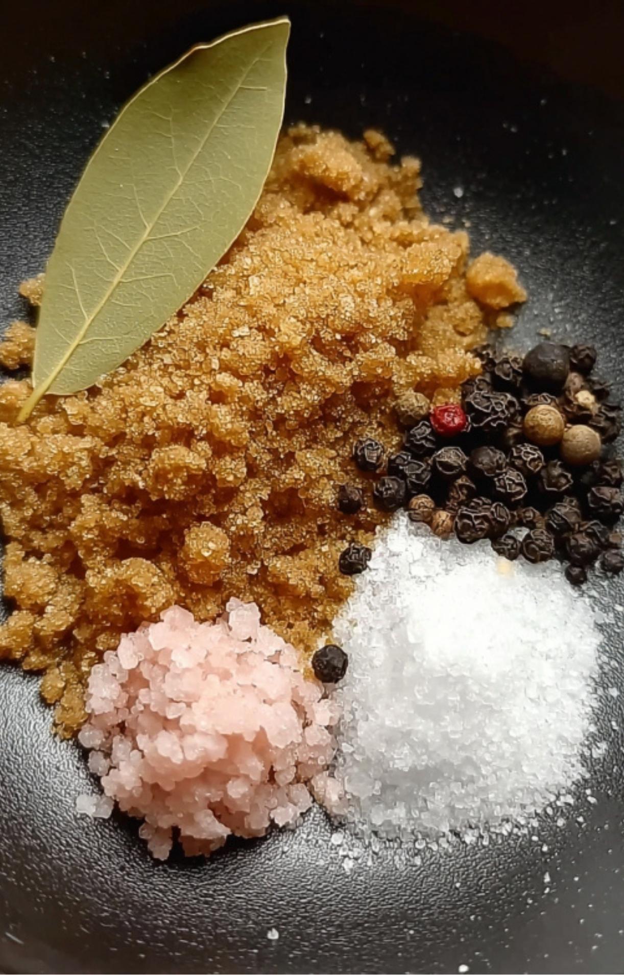 A high-detail shot of coarse sea salt, pink curing salt, black peppercorns, brown sugar, and a single green bay leaf in a black bowl.
