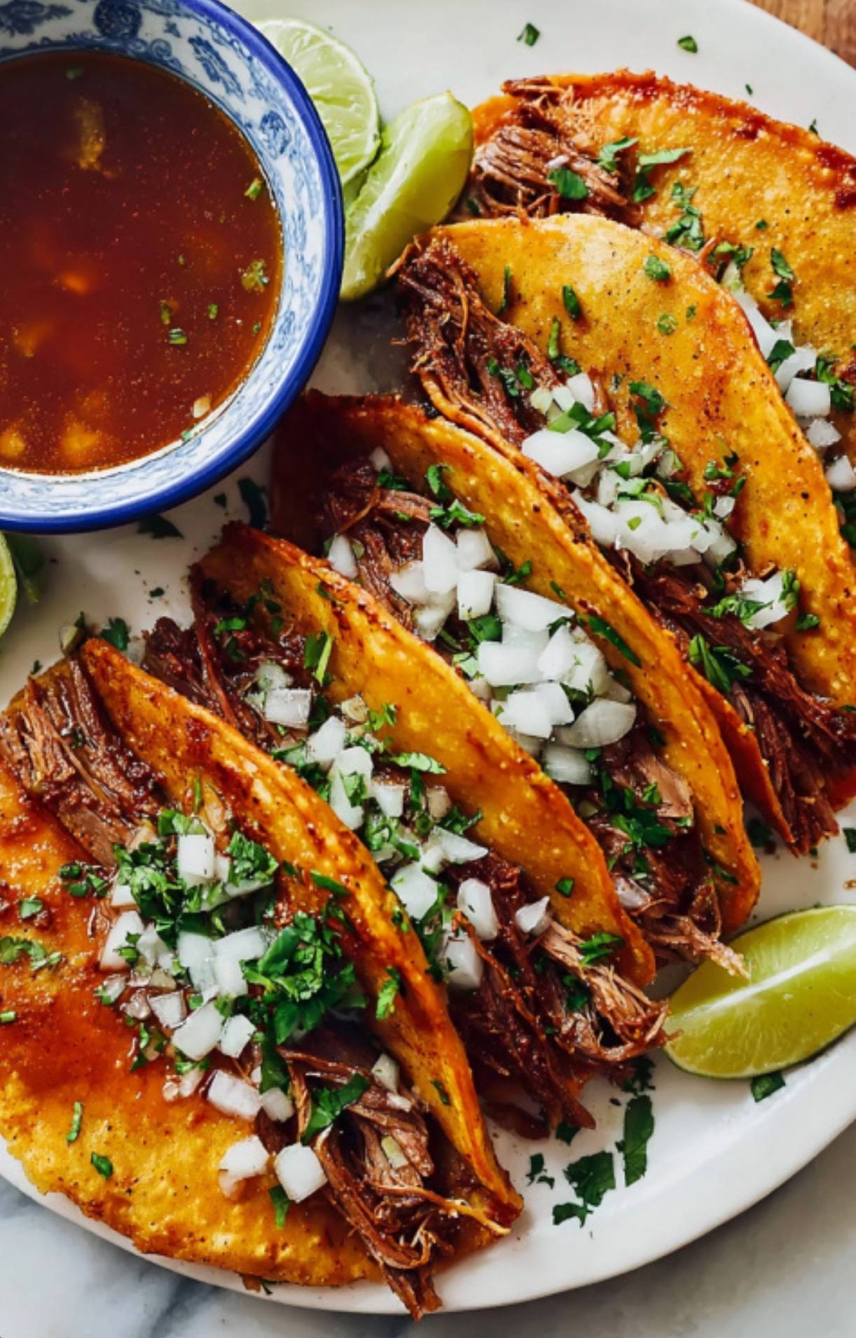 A white plate holding four loaded birria tacos garnished with diced white onions and fresh cilantro, served with lime wedges and a side of dipping sauce.