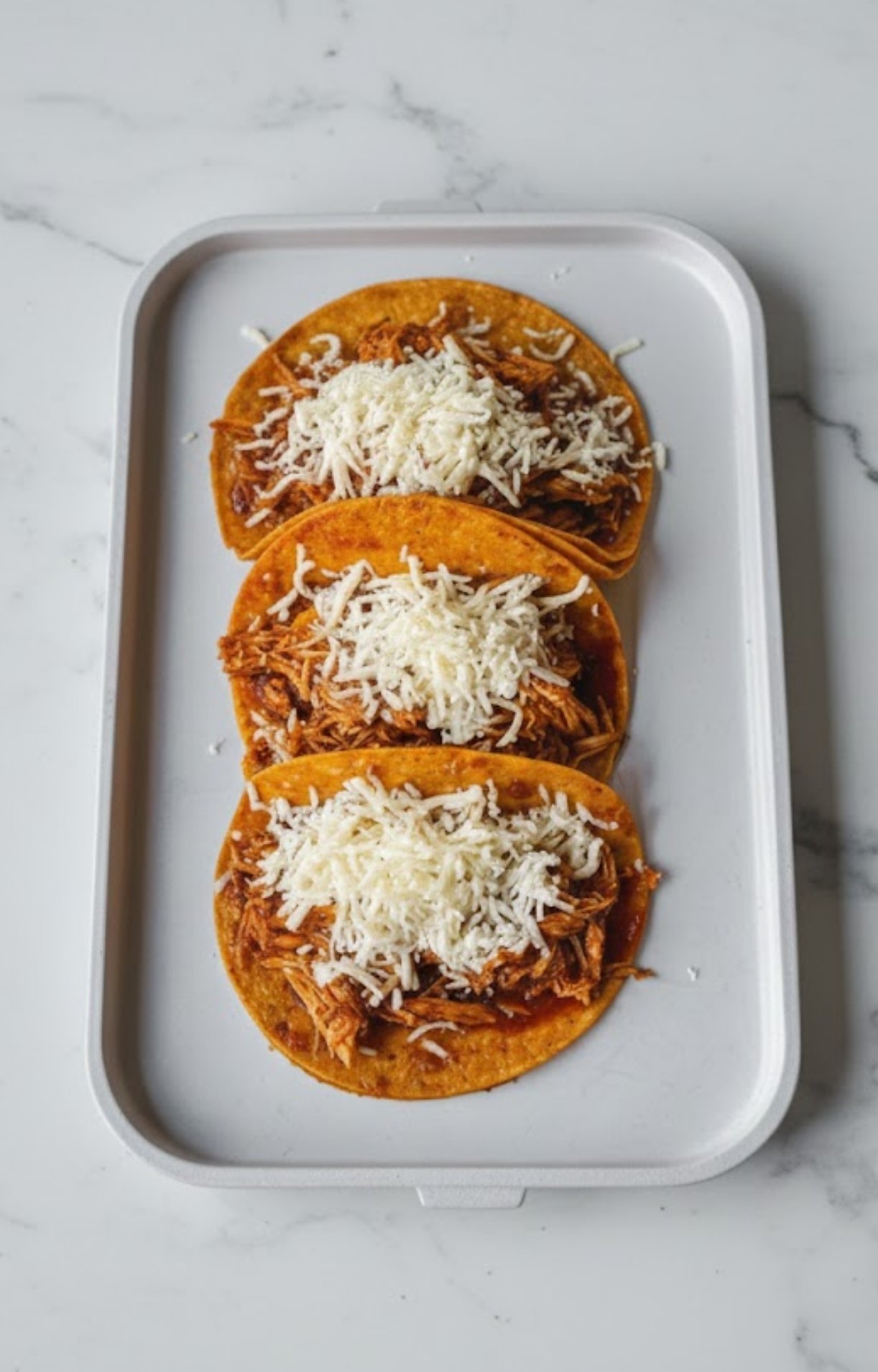 Three dipped tortillas on a tray topped with the saucy shredded chicken and a pile of shredded white cheese.