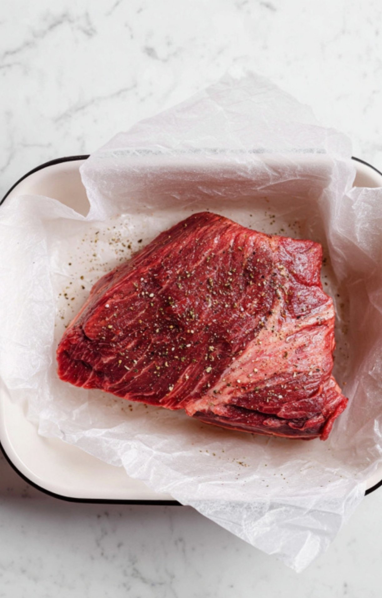 A large cut of raw beef chuck roast sitting in a parchment-lined tray, seasoned generously with salt and black pepper.
