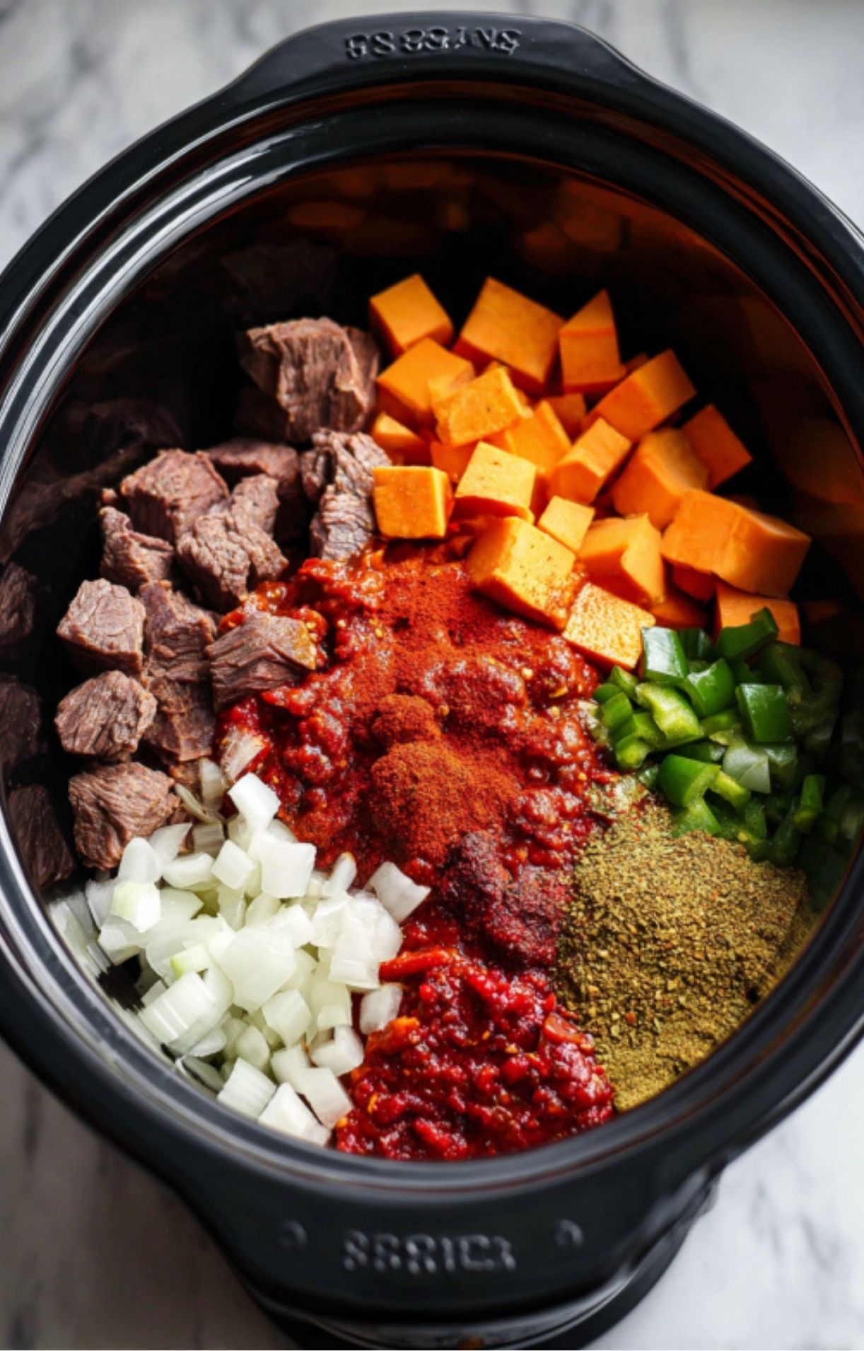Layered raw ingredients including seared beef, diced sweet potatoes, onions, peppers, and tomato base topped with chili spices in a black slow cooker.