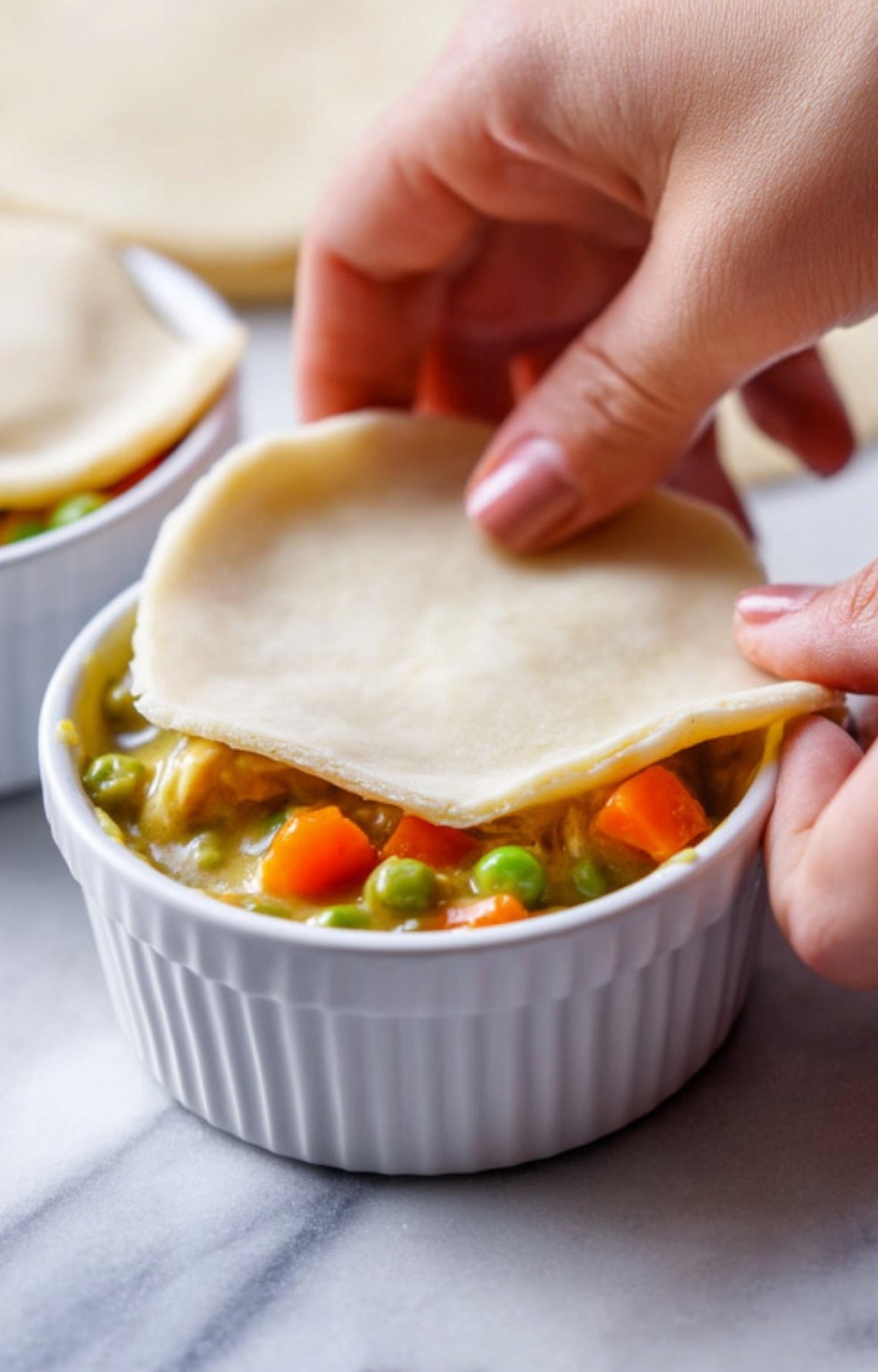 A person's hands carefully placing a round circle of raw puff pastry dough onto a white ramekin filled with turkey pot pie mixture.