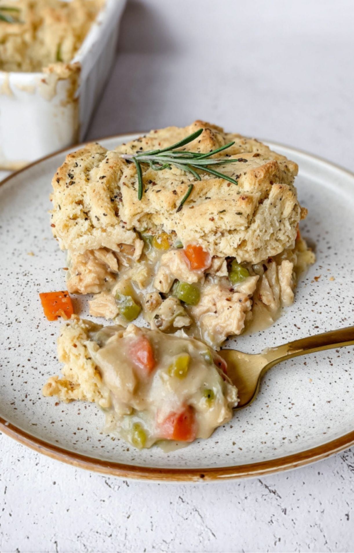 A single serving of turkey pot pie on a speckled plate, showing a fluffy biscuit crust over a creamy vegetable and turkey center, garnished with rosemary.