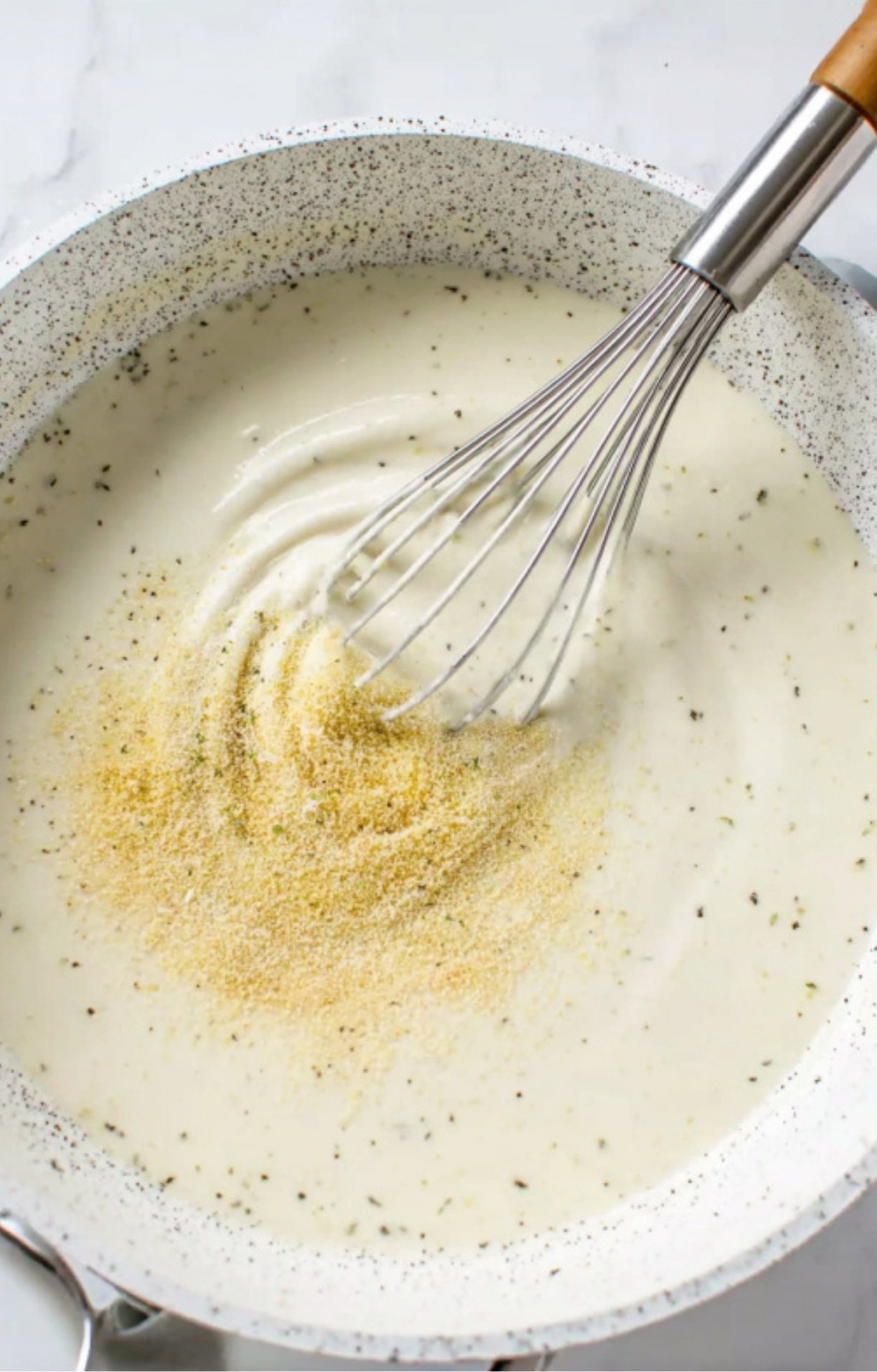 Adding garlic powder and herbs to a creamy white sauce in a saucepan while whisking.