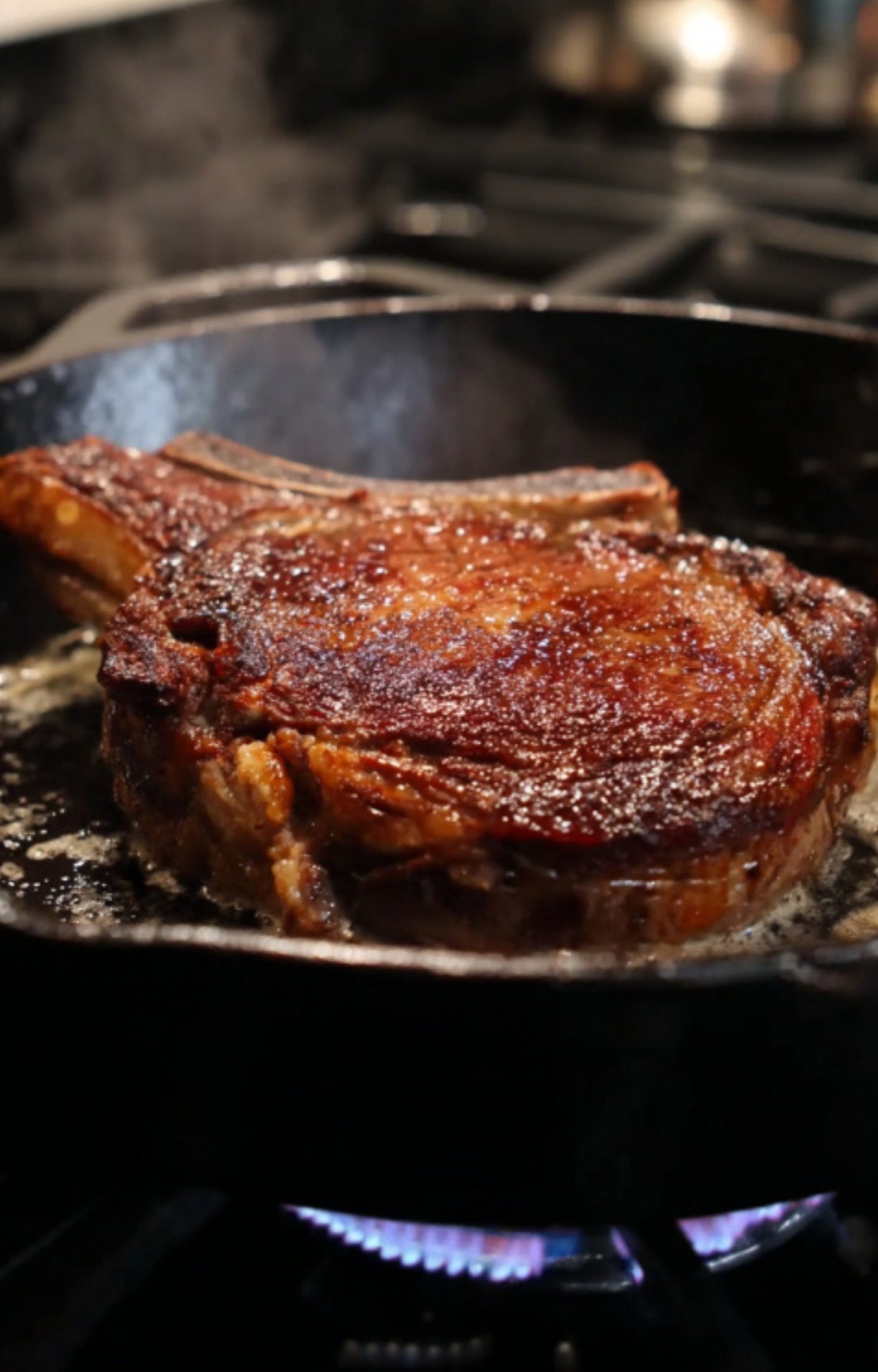 A close-up shot of a rib eye steak developing a deep golden-brown crust while searing over a blue gas flame.