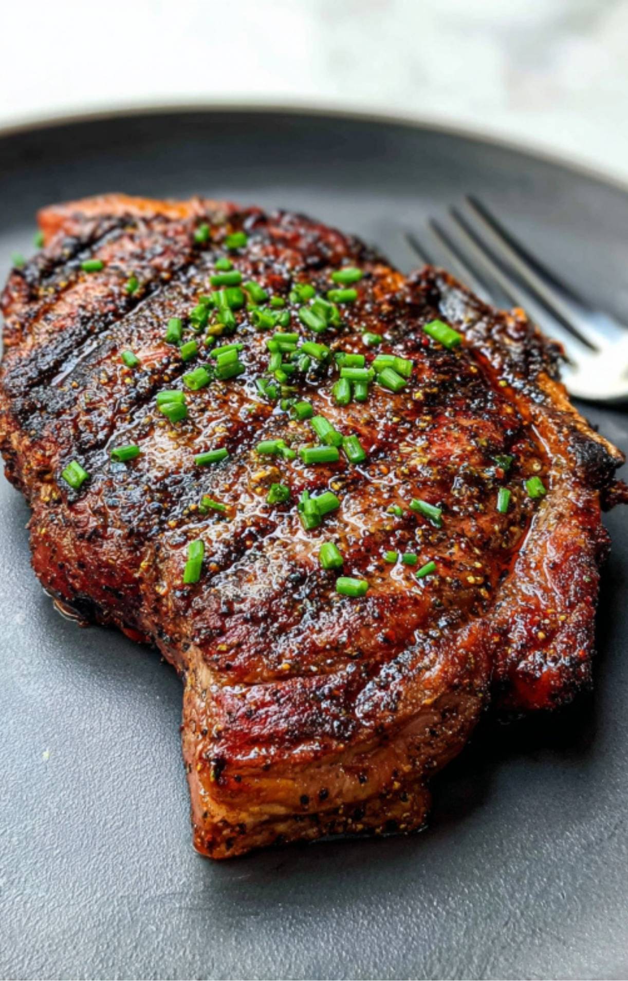 A whole, perfectly grilled ribeye steak resting on a dark plate, garnished with a sprinkle of bright green chives and served with a fork.