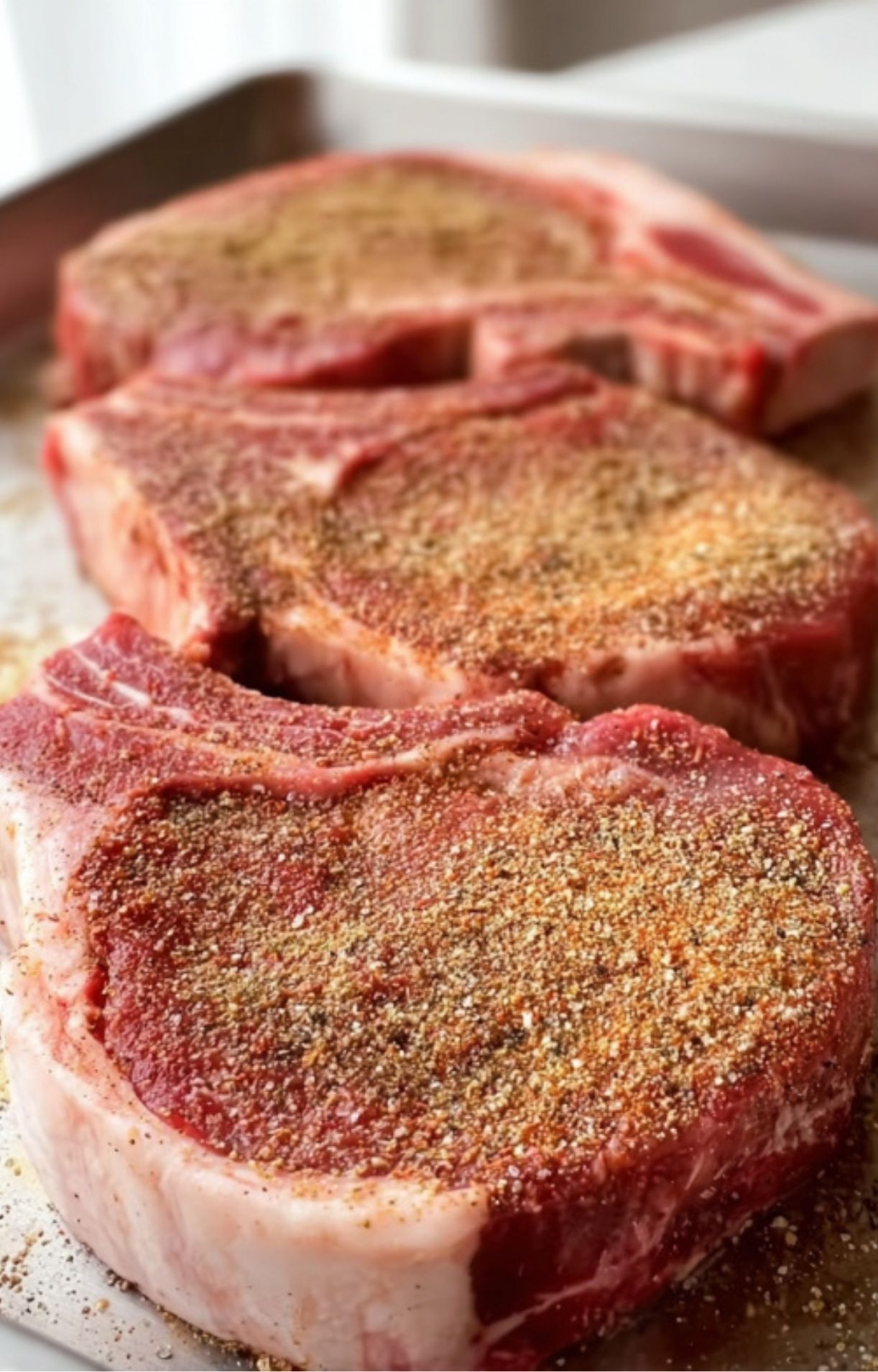 A close-up of raw bone-in rib eye steaks being generously coated with a dry rub of salt, pepper, and garlic powder on a metal tray.