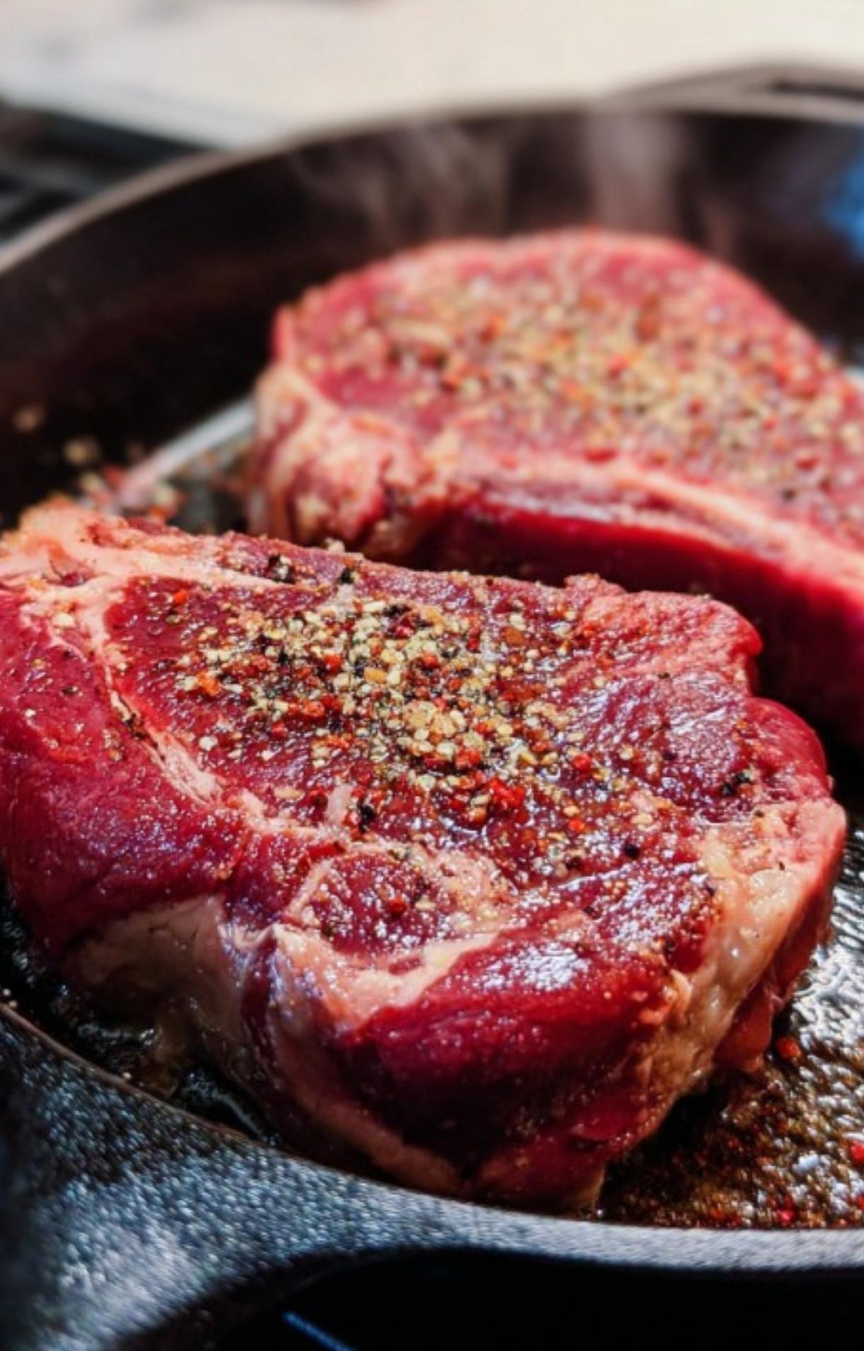 Two thick cuts of rib eye steak seasoned with cracked black pepper and sea salt searing in a hot cast iron skillet.