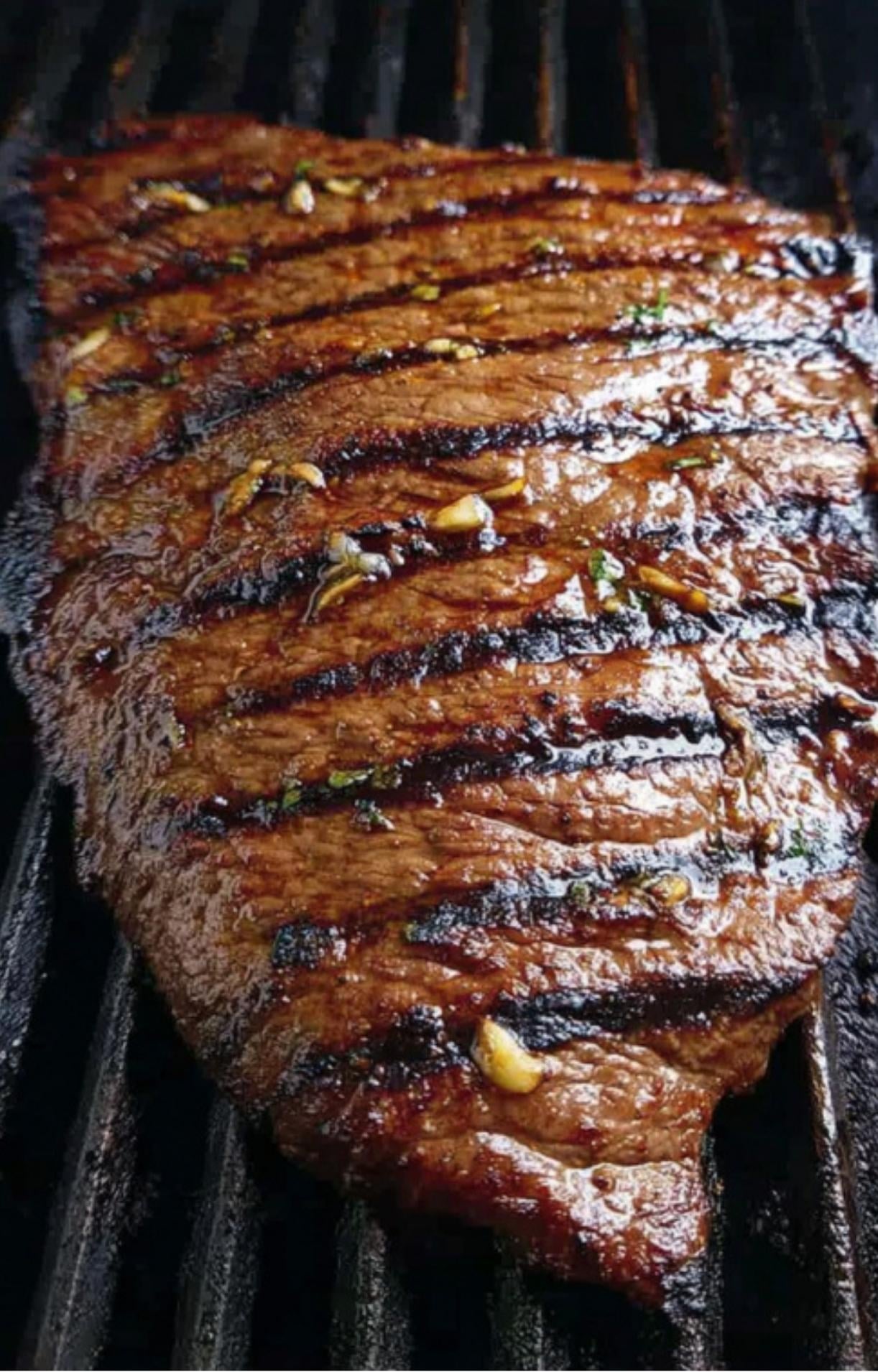 Juicy marinated flank steak sizzling on a grill with charred grill marks and bits of garlic, perfect for summer grilling.