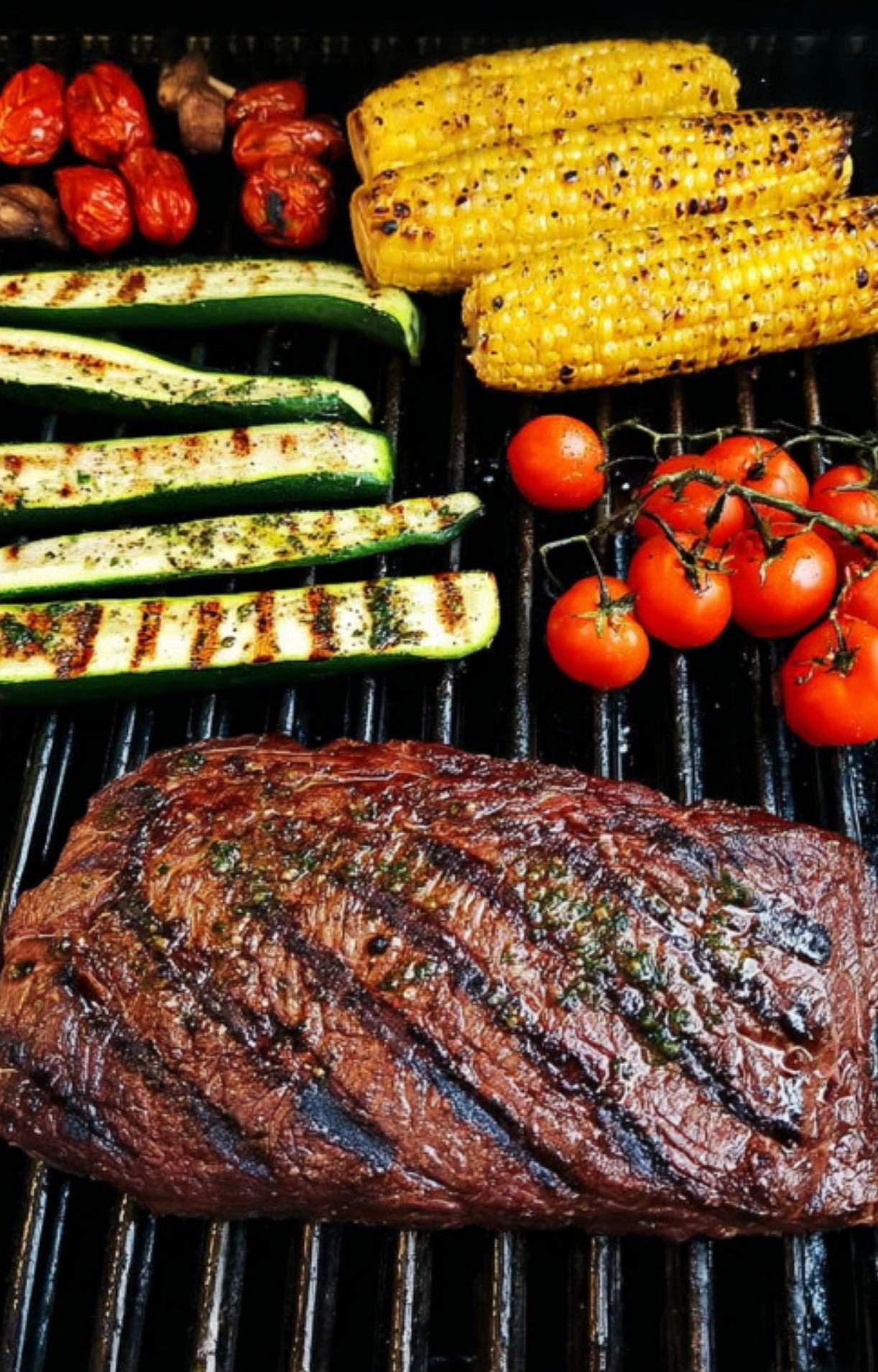 Grilled flank steak and vegetables including corn, zucchini, and cherry tomatoes cooking on a barbecue