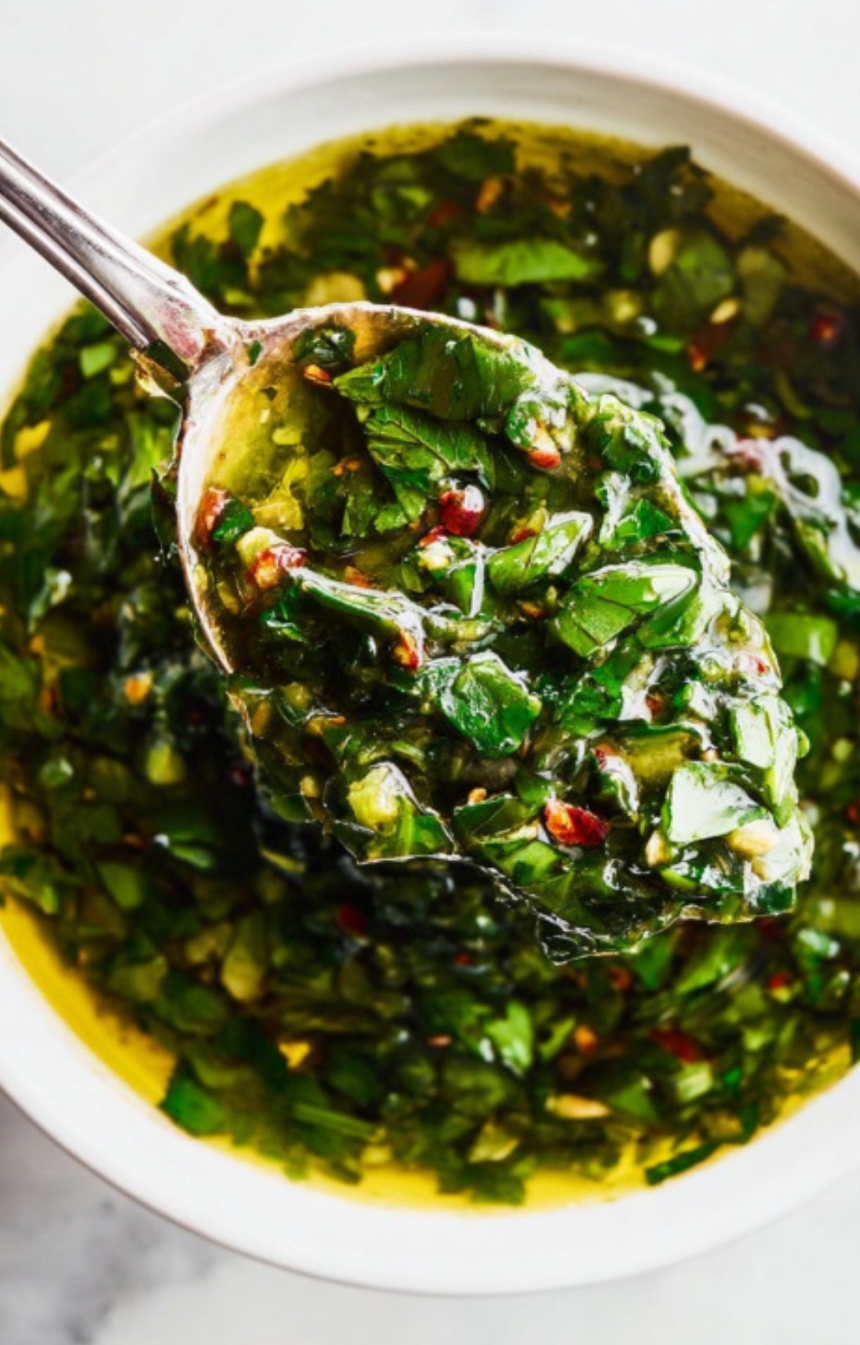 Silver spoon scooping up chunky homemade chimichurri sauce made with parsley, garlic, olive oil, and chili flakes, shown over a white bowl.