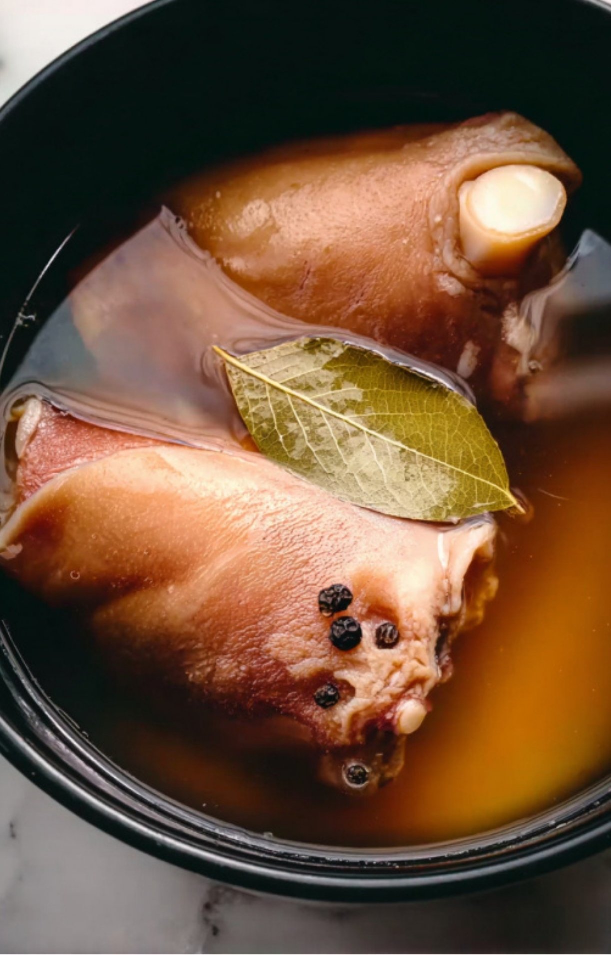 Two pork hocks submerged in a dark liquid brine with peppercorns and a bay leaf, soaking up flavors before hitting the smoker.