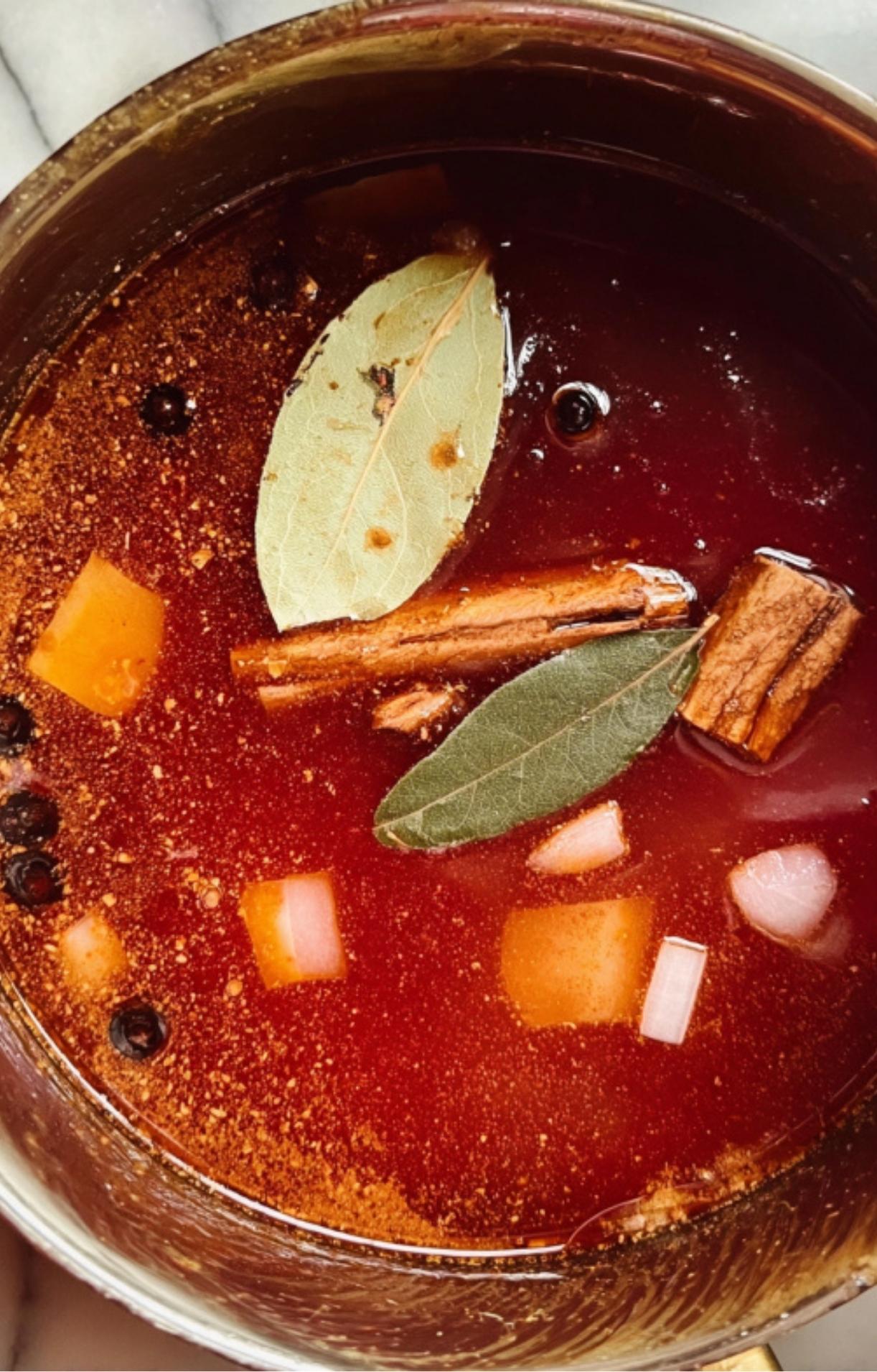 A pot of deep red birria consommé containing bay leaves, cinnamon sticks, and whole peppercorns.