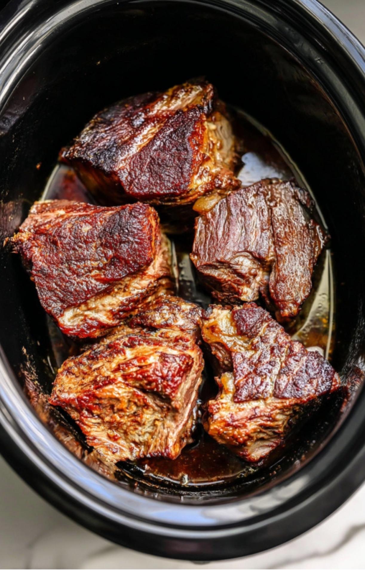 Large pieces of browned, seared beef chuck roast sitting at the bottom of a black slow cooker for Crockpot Birria Tacos.
