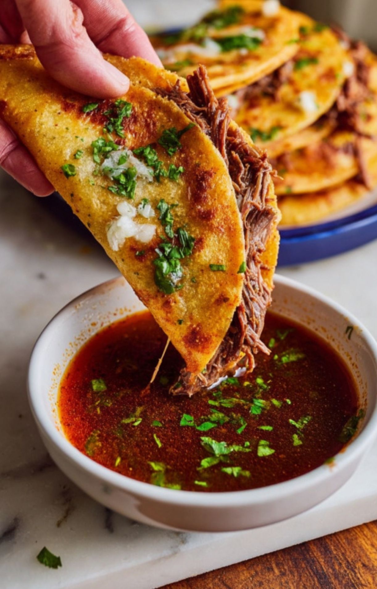 A hand holding a crispy beef birria taco stuffed with shredded meat, onions, and cilantro, dipping the edge into a small bowl of savory consommé.