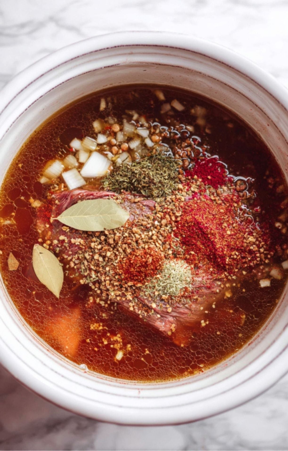 Raw beef submerged in a liquid broth within a white pot, topped with dried bay leaves, chili powder, oregano, and diced onions.