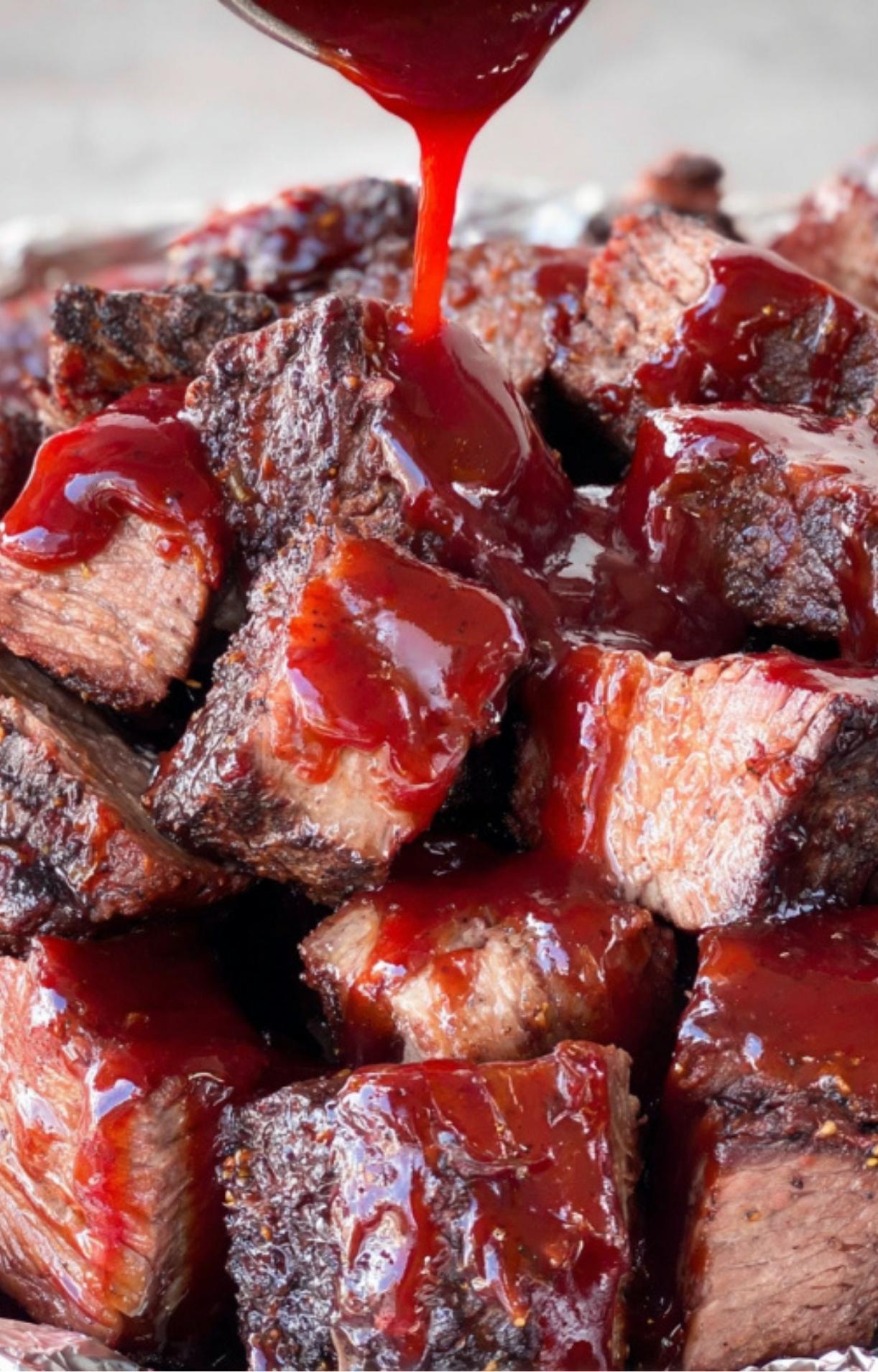 A stream of thick, vibrant red barbecue sauce being poured over a pile of smoked beef cubes.
