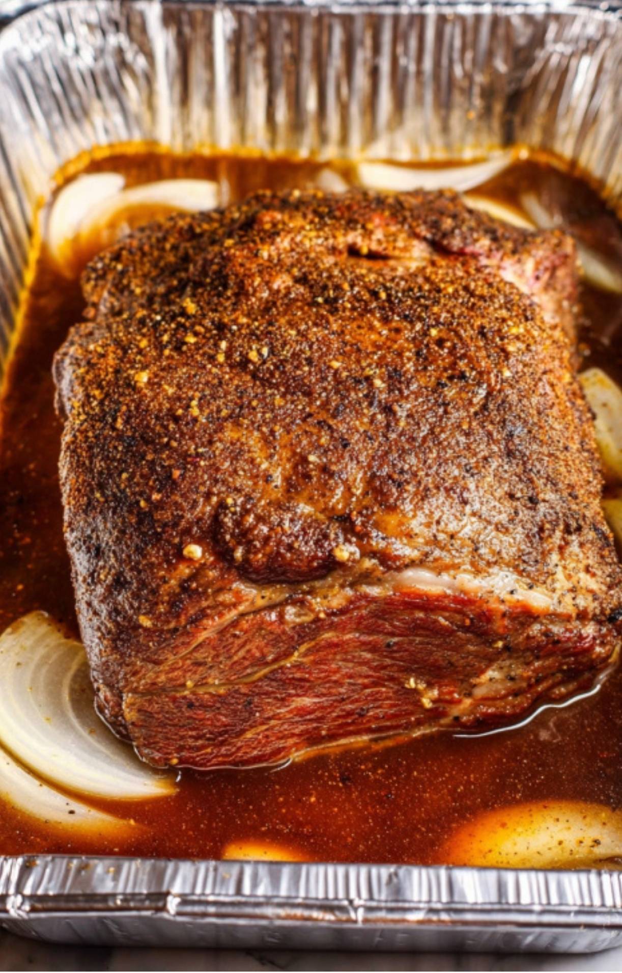 The smoked beef placed in an aluminum pan with onions and savory liquid to finish cooking until tender.