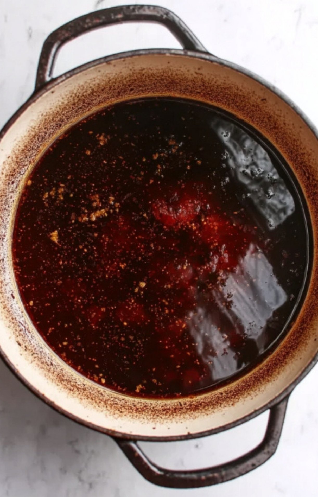 A top-down view of a dark red wine and broth mixture simmering in a black Dutch oven to scrape up browned bits.
