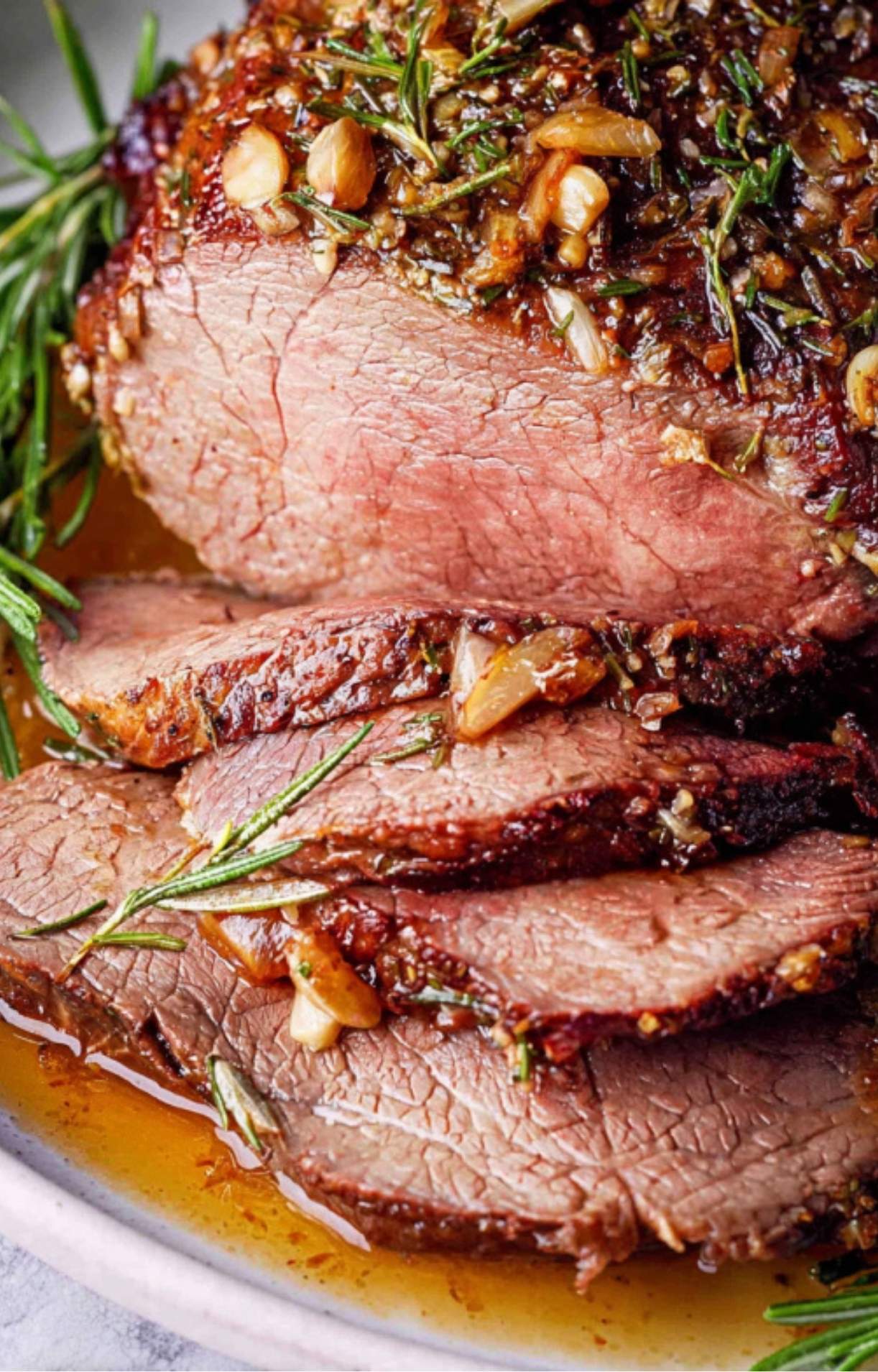 A detailed shot of tender, pink Chuck Tender Roast slices topped with roasted garlic and herbs in a pool of pan juices.