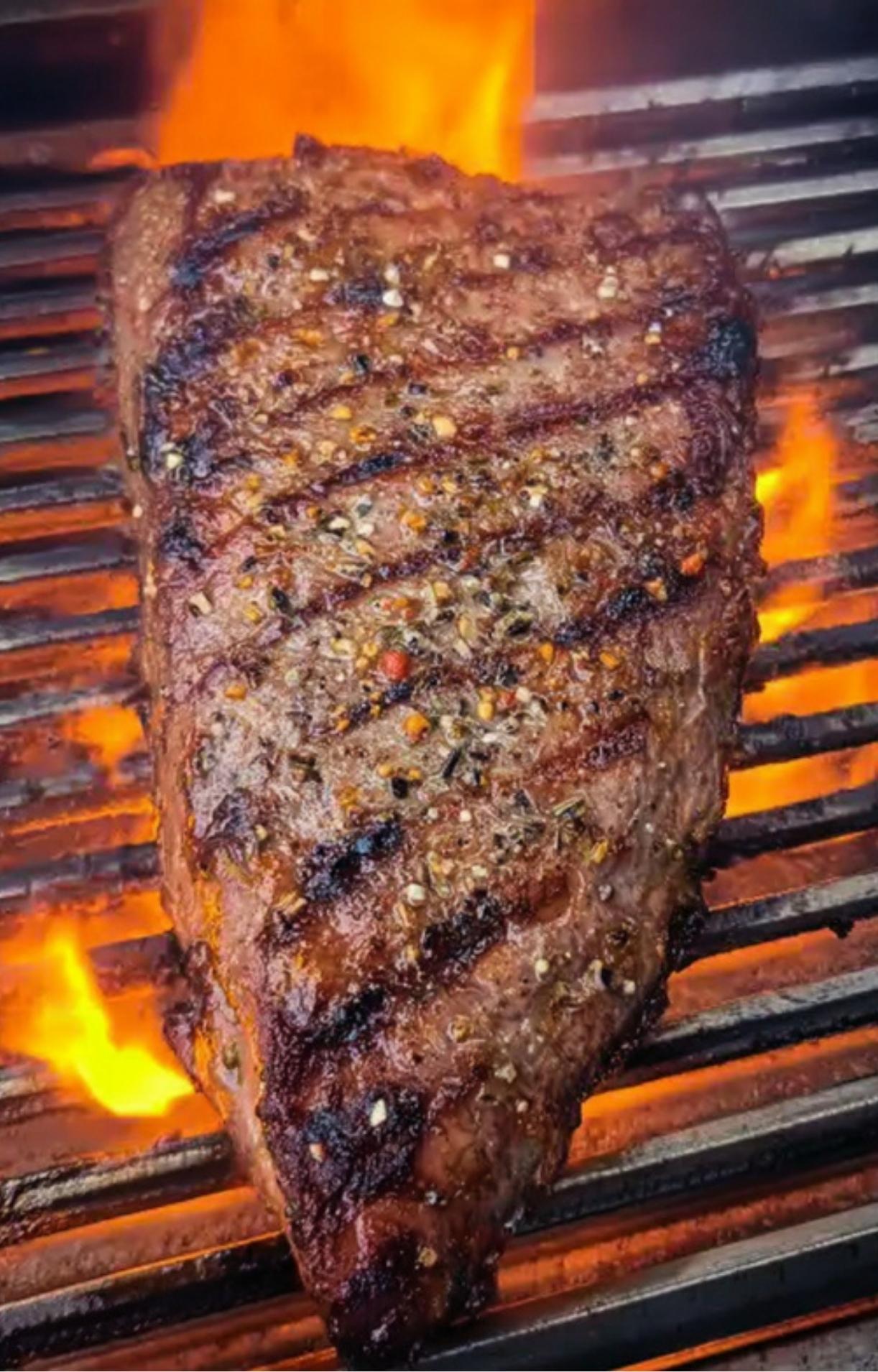 Seasoned tri tip steak cooking on a hot grill with orange flames, showing sear marks.