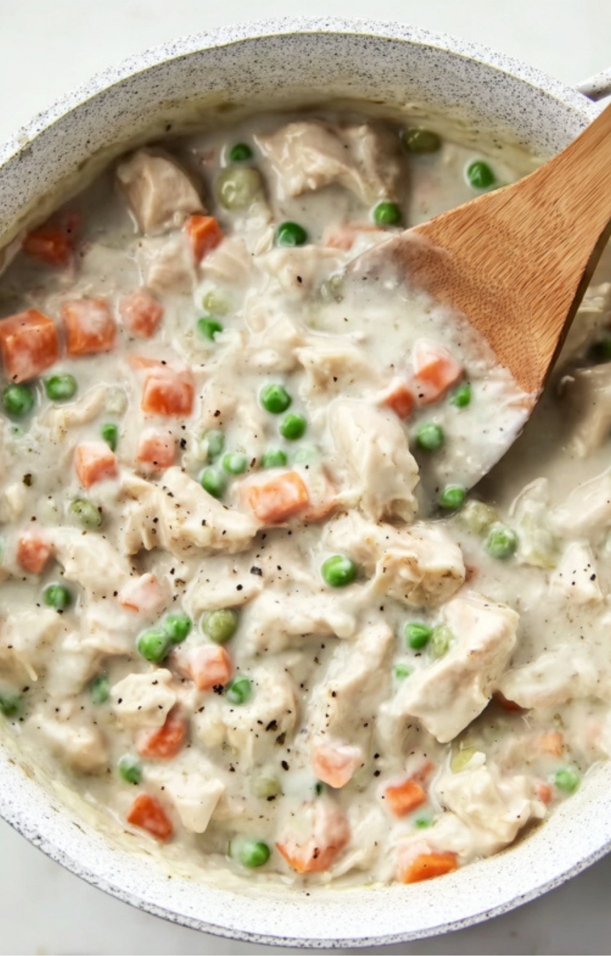 A wooden spoon stirring shredded turkey, green peas, and diced carrots into a thick, creamy white sauce in a pot.